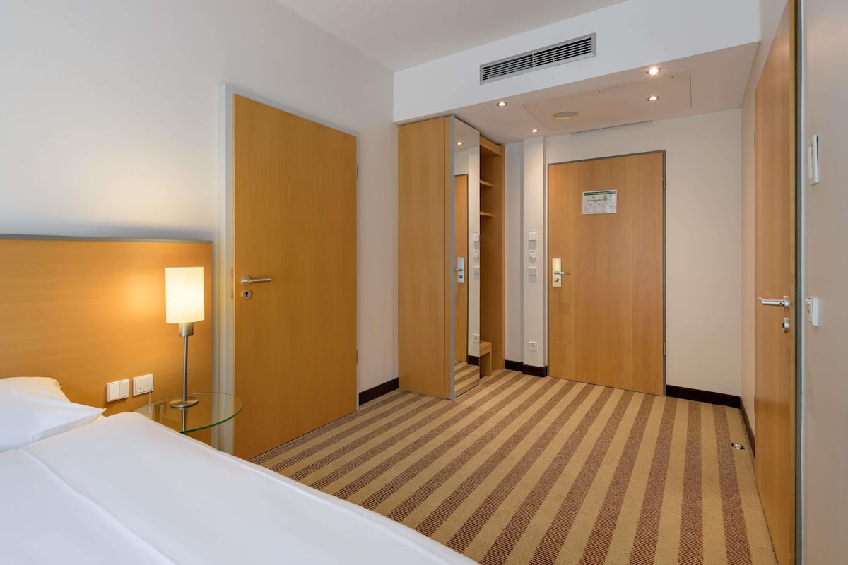 A hotel room with a bed, a nightstand, a lamp, a mirror, and a door leading to a bathroom.