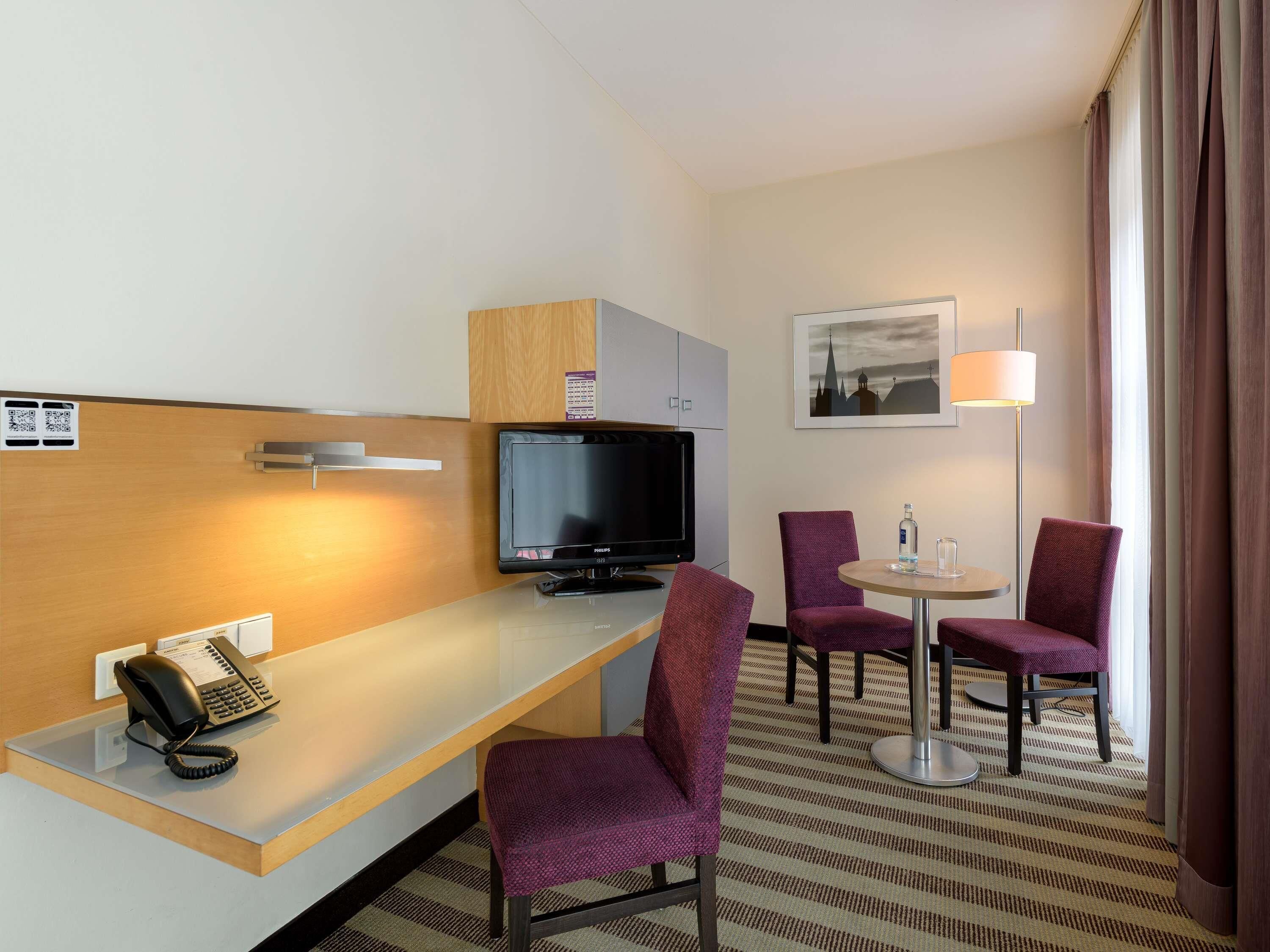 Habitación de hotel con escritorio, televisor de pantalla plana, dos sillas moradas, una mesita de noche con lámpara y una ventana con cortinas.
