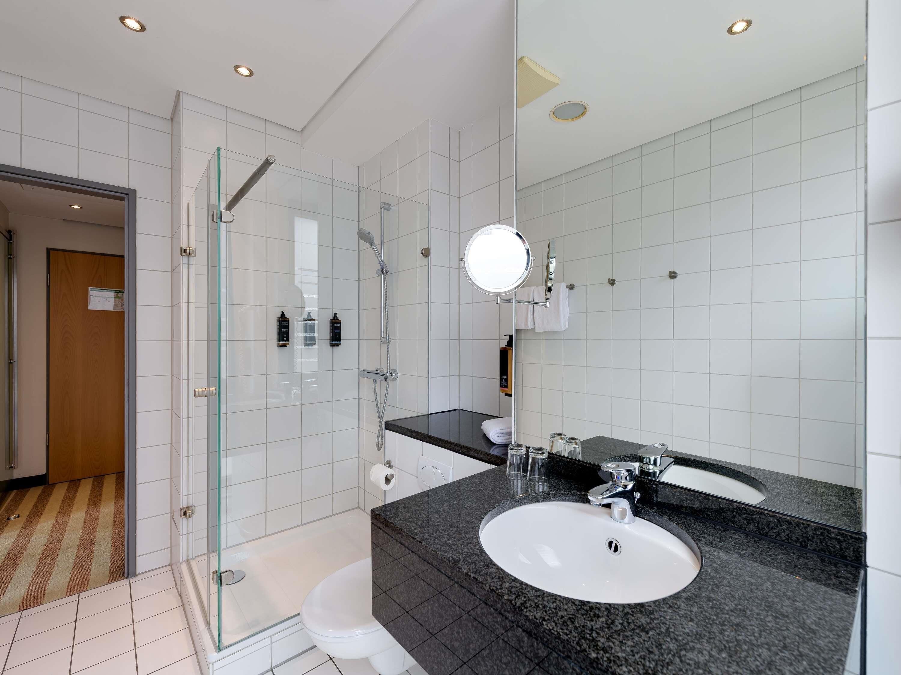 A modern bathroom with a glass shower enclosure, a white toilet, a sink with a granite countertop, and a mirror.