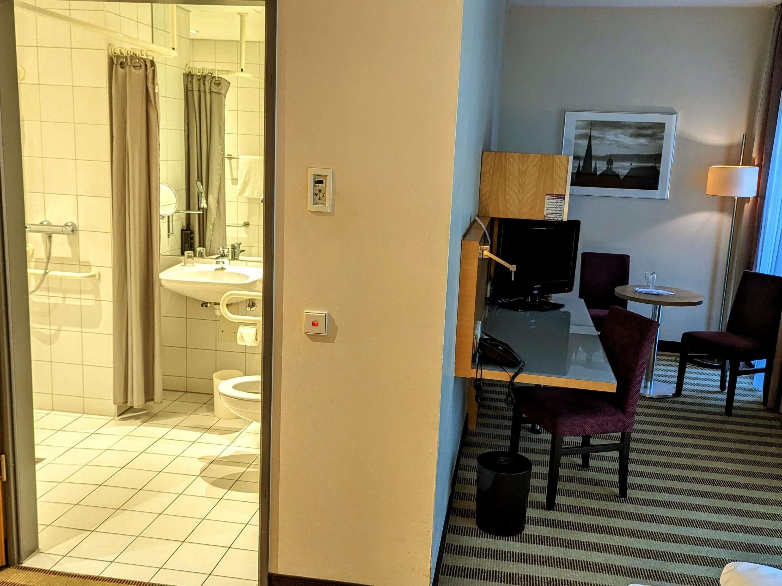A hotel room with a bathroom, a desk with a computer, two chairs, a small table, and a lamp.