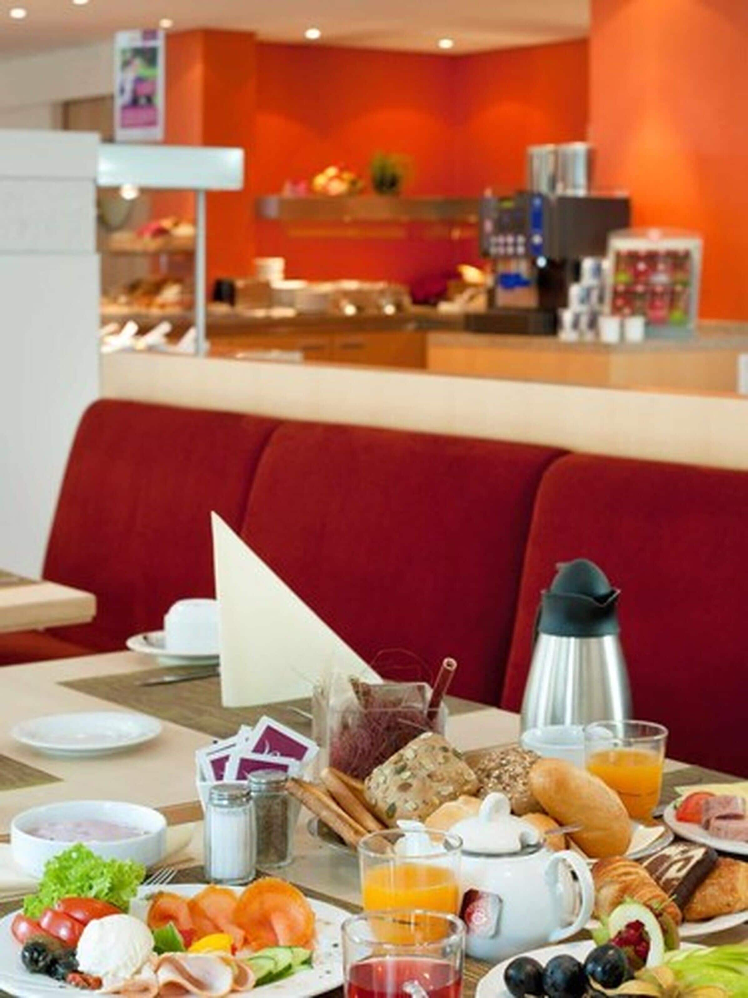 A breakfast buffet with a variety of foods, including fruits, pastries, and beverages, set in a modern dining area with a red booth.