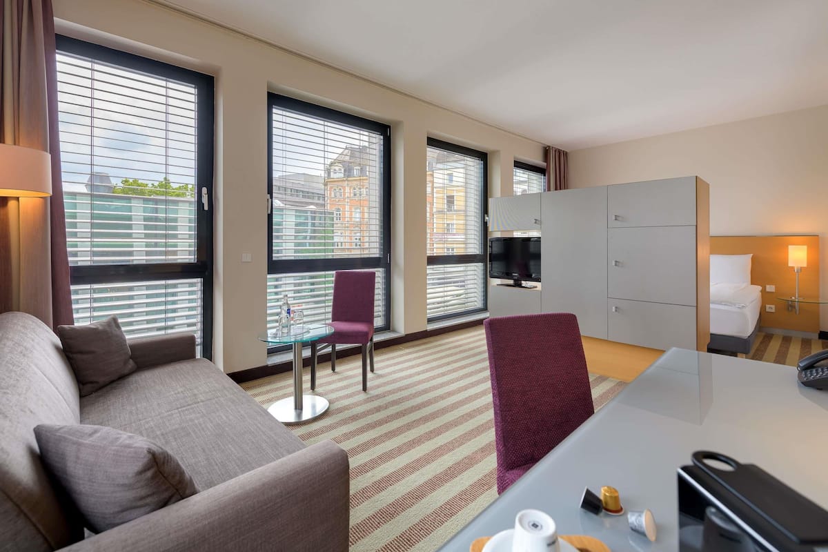 A hotel room with a sofa, a dining table, a TV, and a minibar.