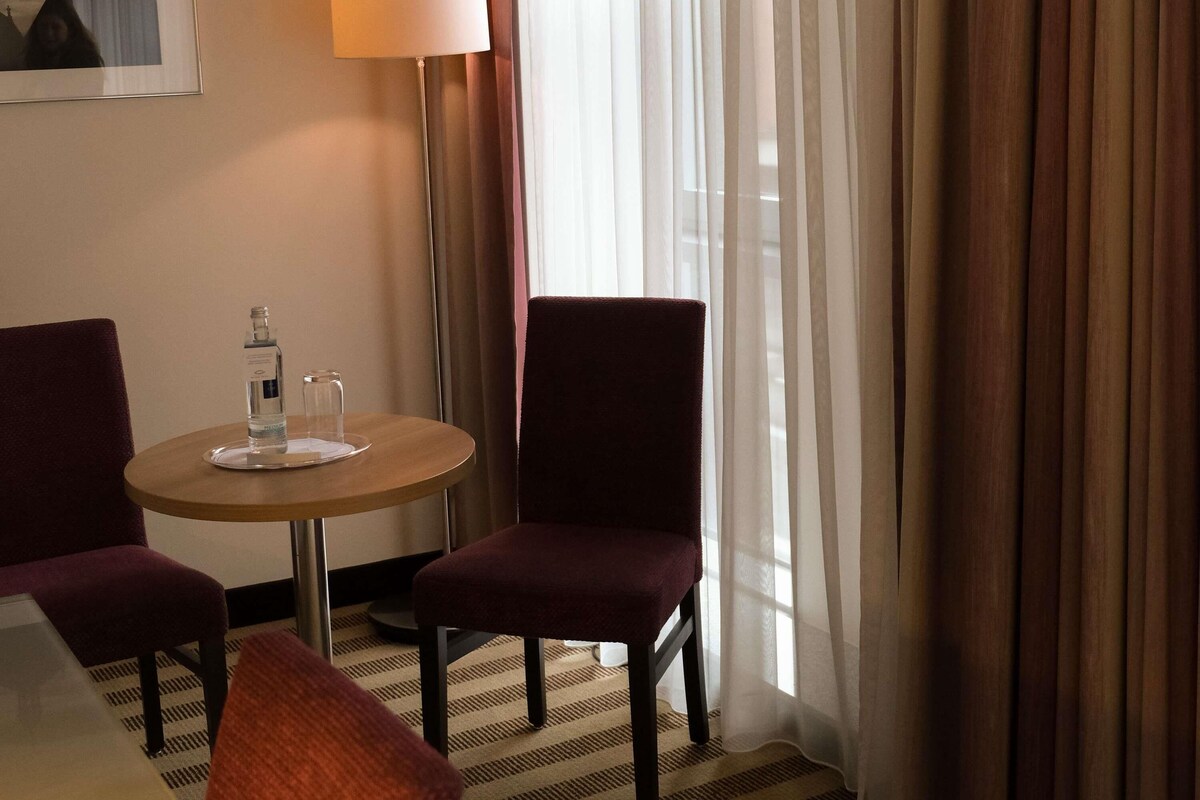 A hotel room with a round table, two chairs, a lamp, and a window with curtains.