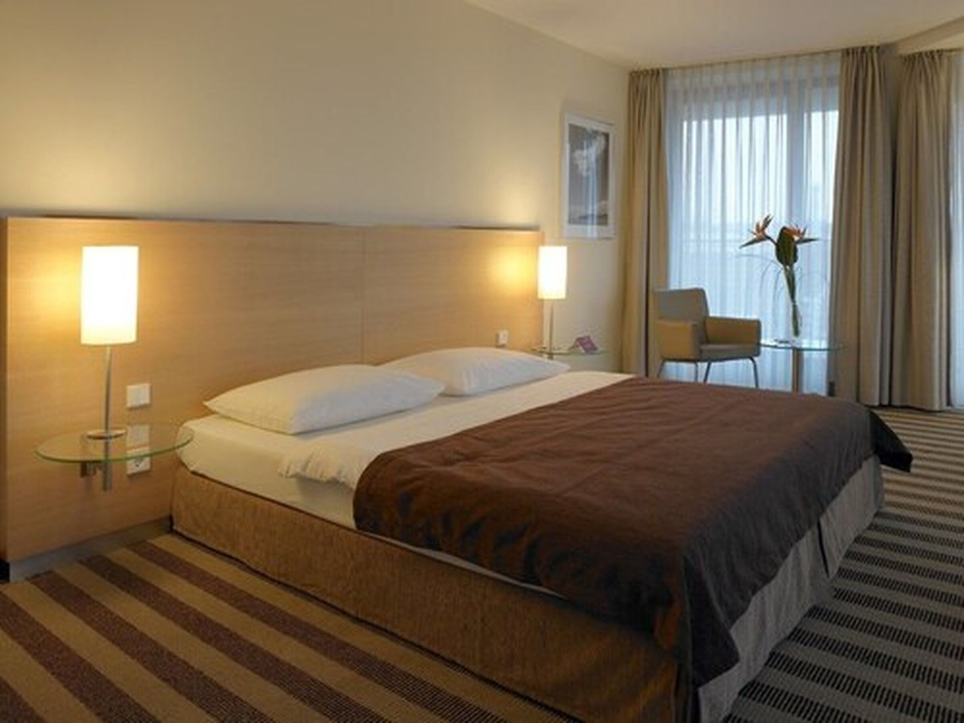 A hotel room with a large bed, two bedside lamps, a small table with a chair, and a window with curtains.