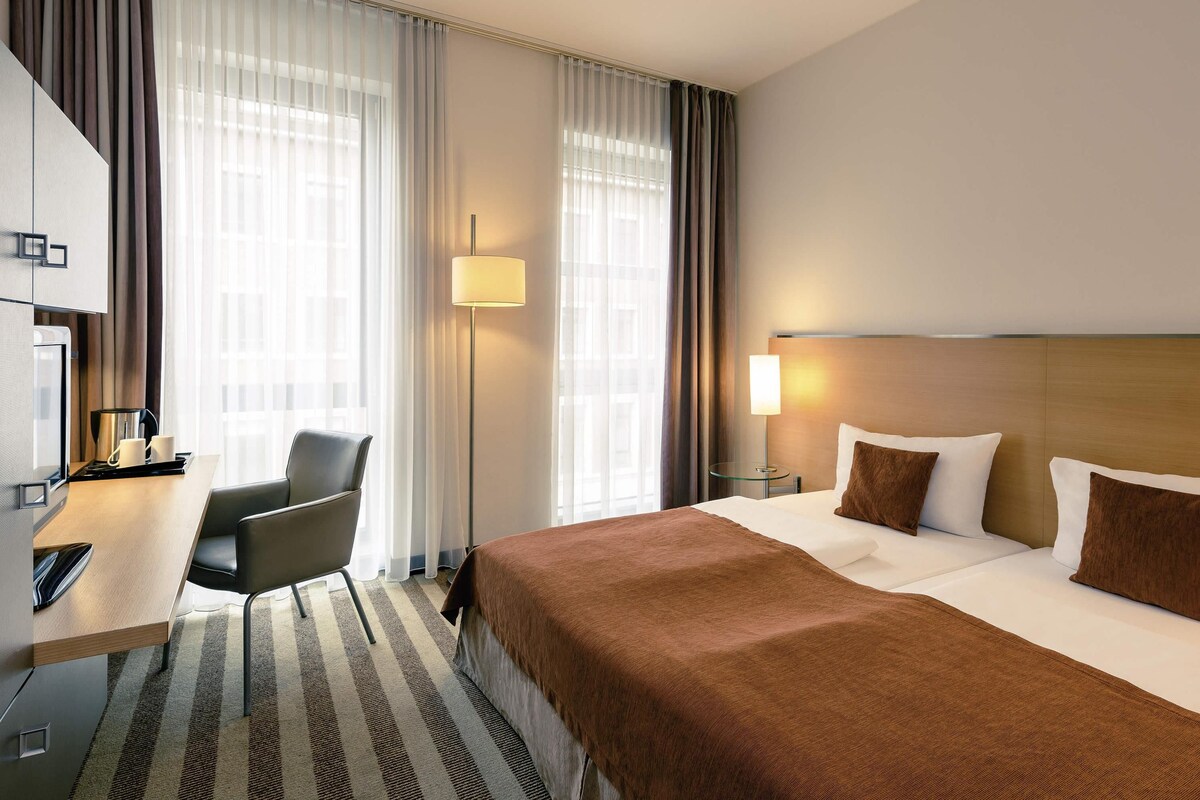 A hotel room with a bed, a desk with a chair, a lamp, and a TV.
