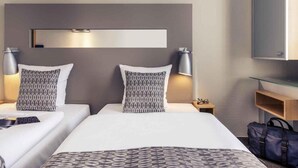 A hotel room with two beds, a bedside table with a lamp, a small nightstand, a wardrobe, and a mirror on the wall.