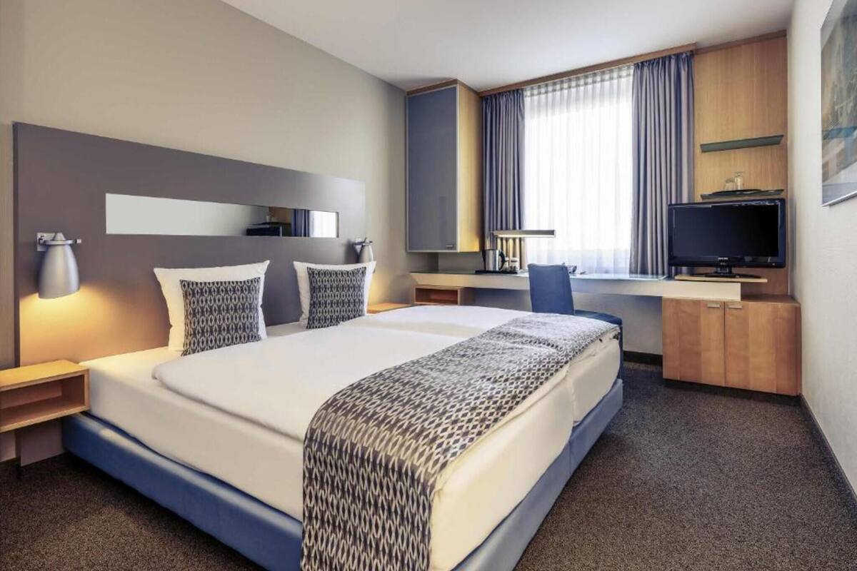 A hotel room with a large bed, a desk with a computer, a TV, and a window with curtains.