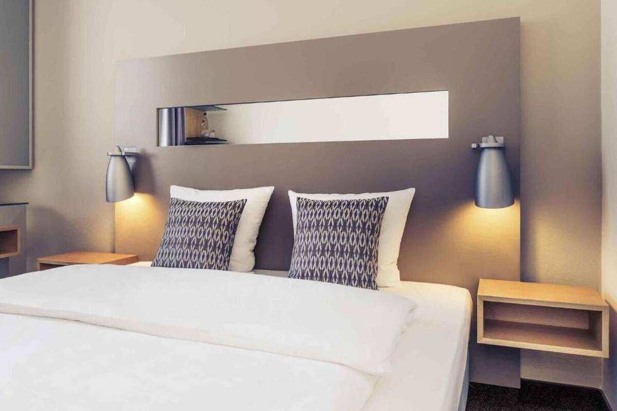 A hotel room with a bed, two bedside tables, wall-mounted lamps, and a shelf above the bed.