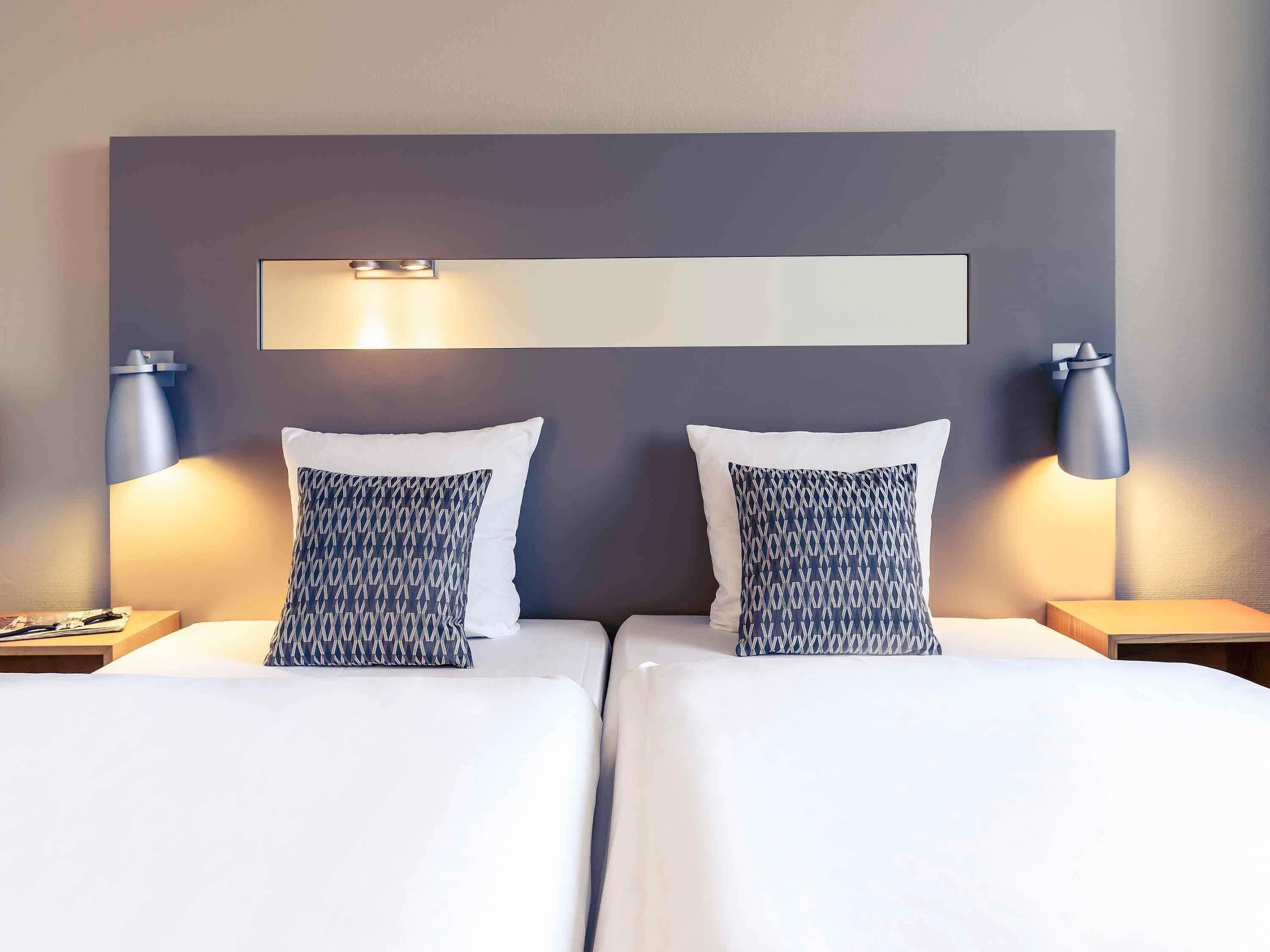 Un lit double avec deux oreillers, une table de chevet, une lampe fixée au mur et une petite table de nuit.