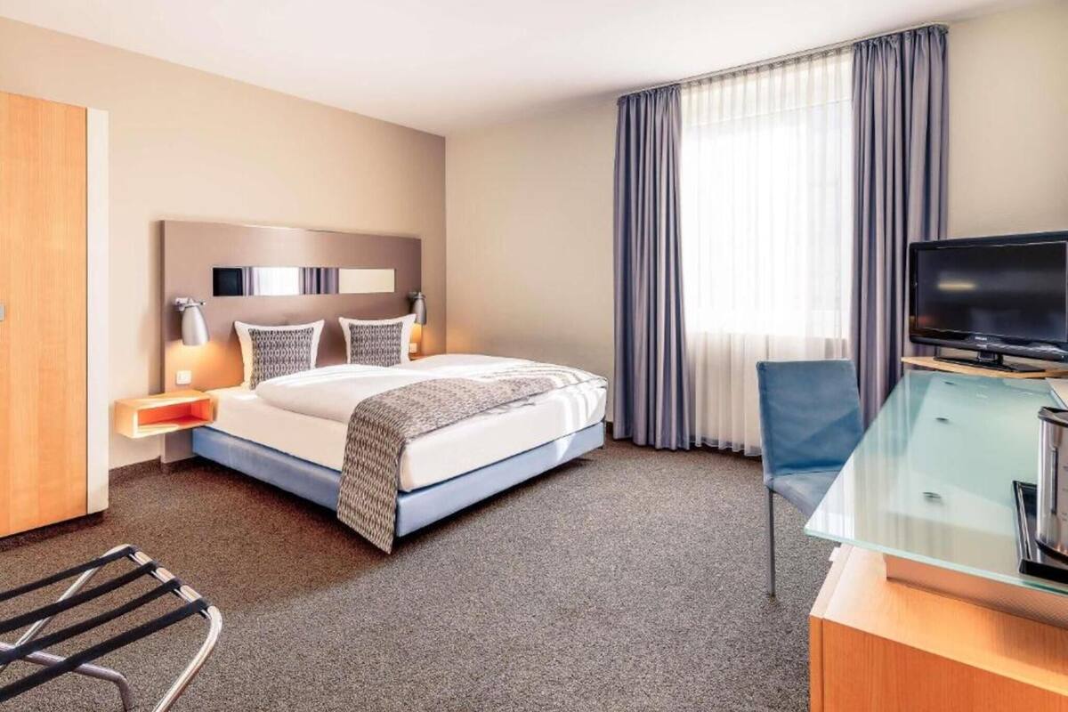 A hotel room with a bed, a desk with a television, a chair, and a window with curtains.