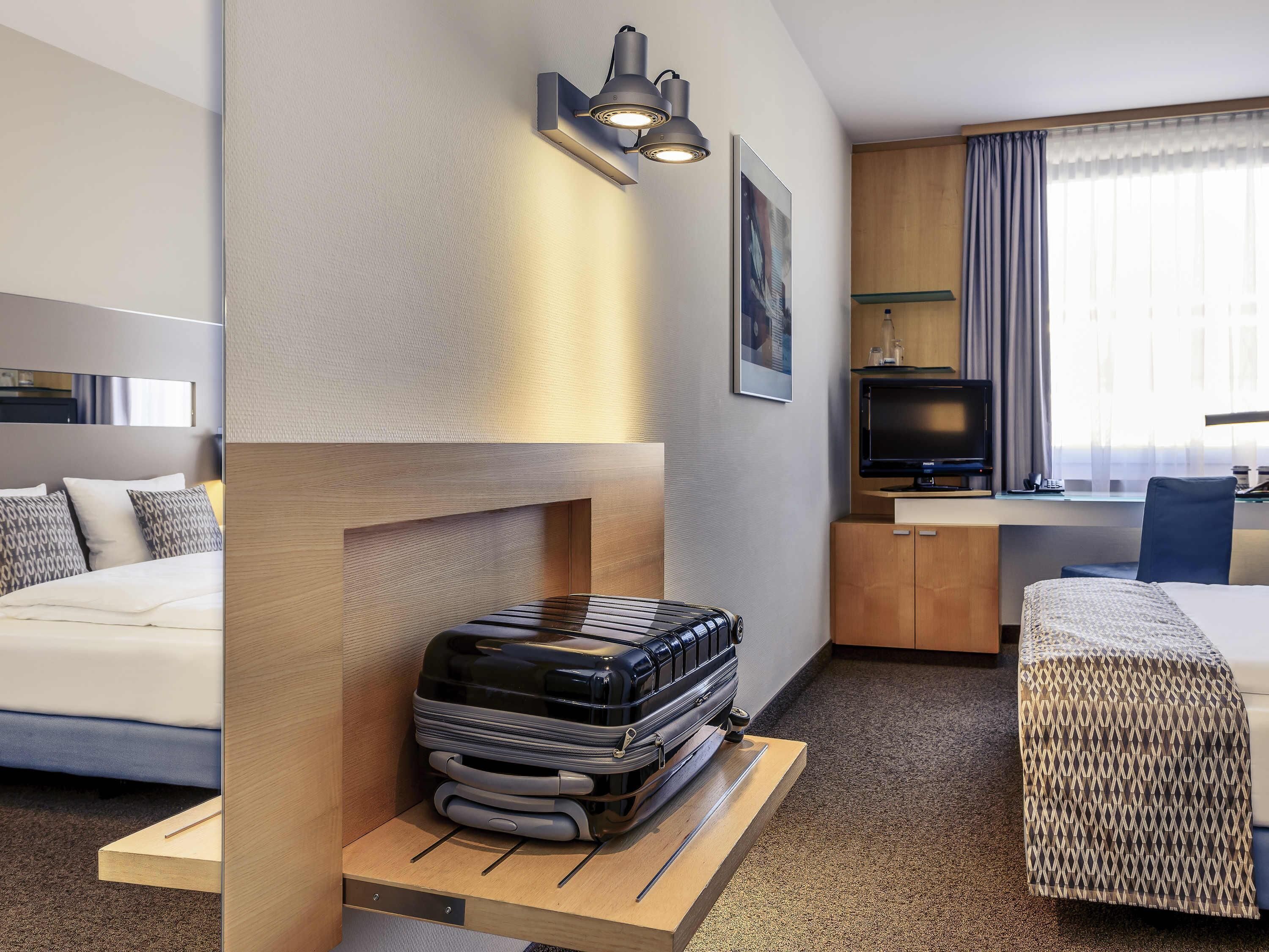 Una habitación de hotel con cama, escritorio, televisor y lámpara fijada a la pared.