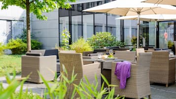 An outdoor seating area with wicker furniture and umbrellas, surrounded by greenery and a modern building facade.