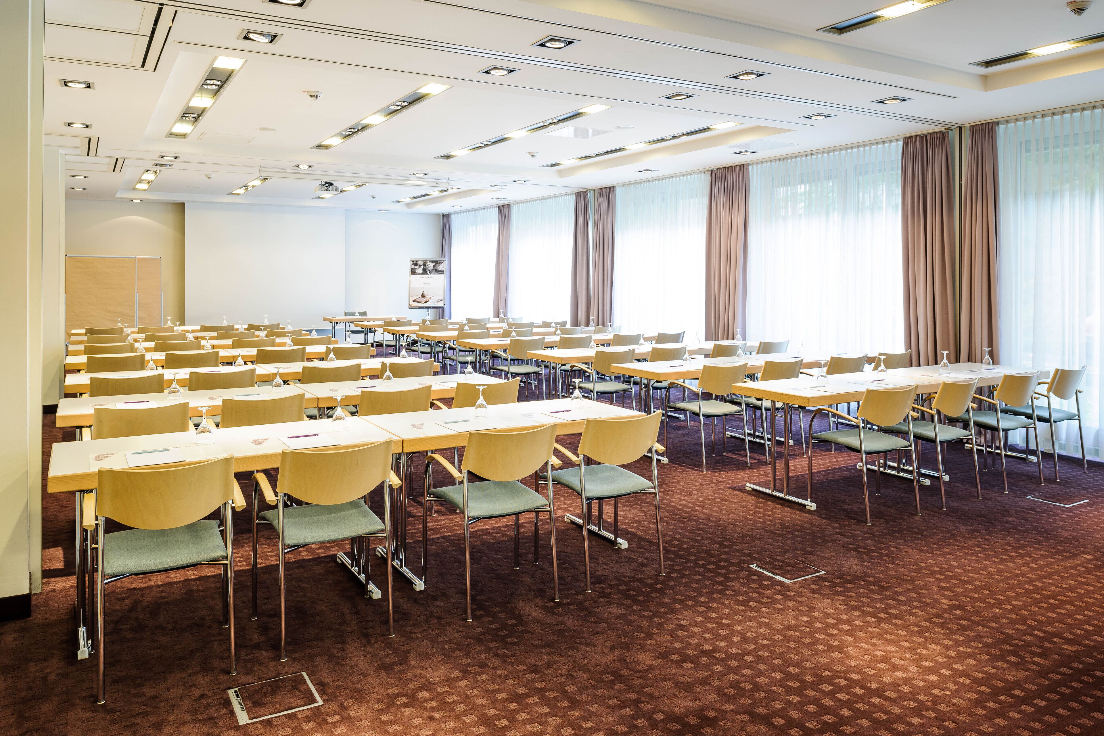 Una sala conferenze con file di tavoli e sedie, ampie finestre con tende e pavimento con moquette.