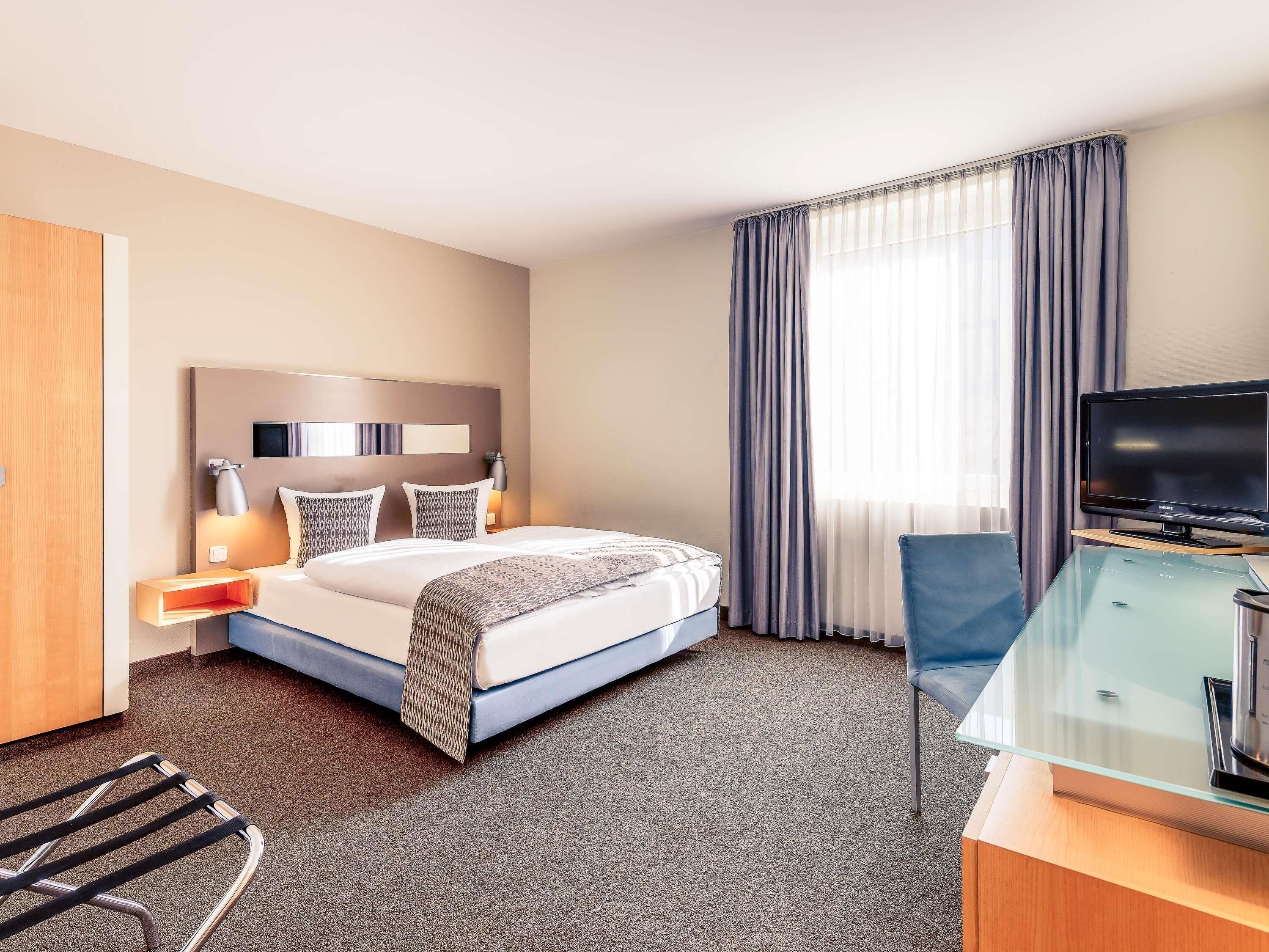 A hotel room with a bed, a desk with a television, a chair, and a window with curtains.