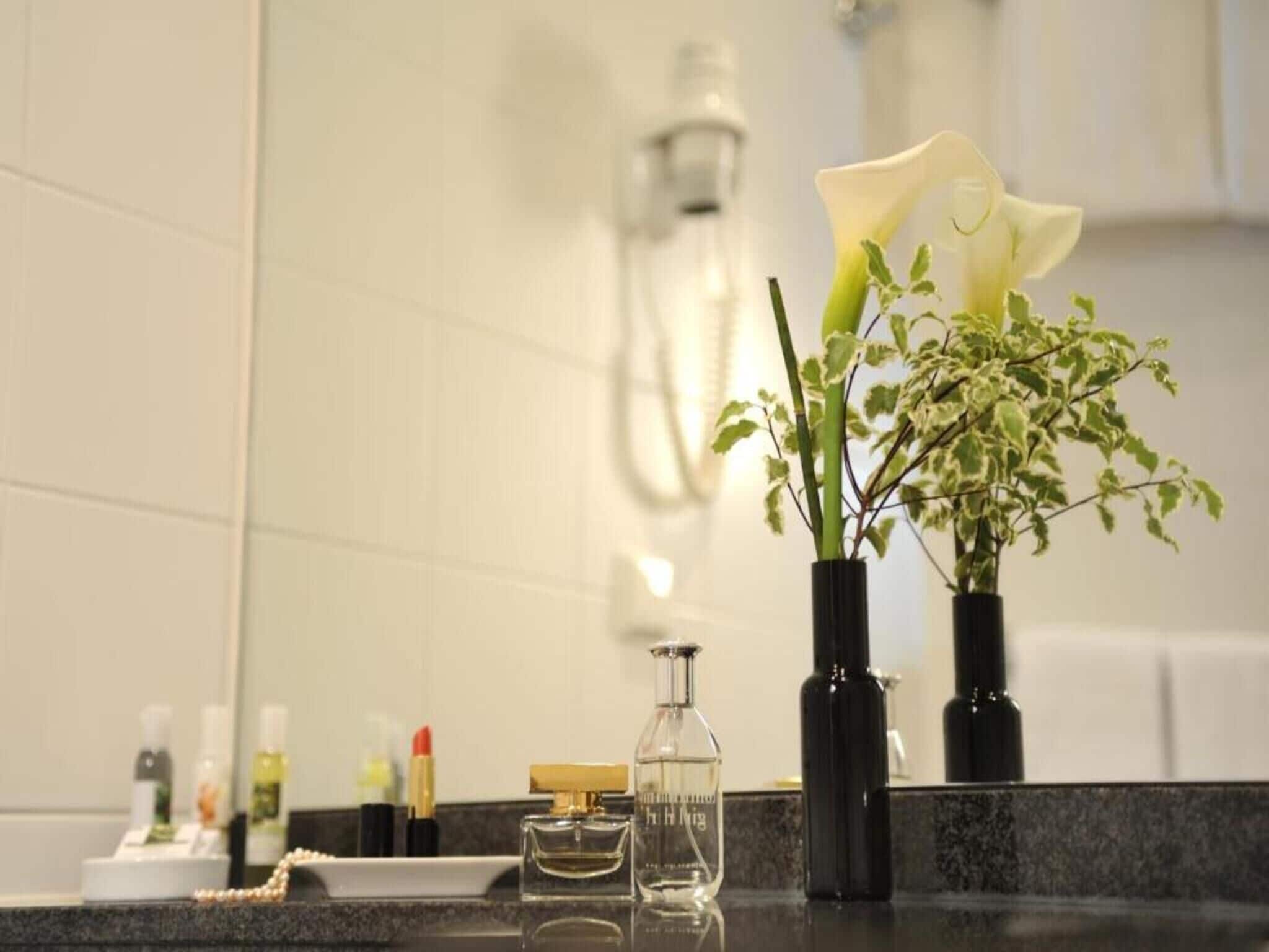 A bathroom with a mirror, a vase with a white flower, and toiletries on the counter.