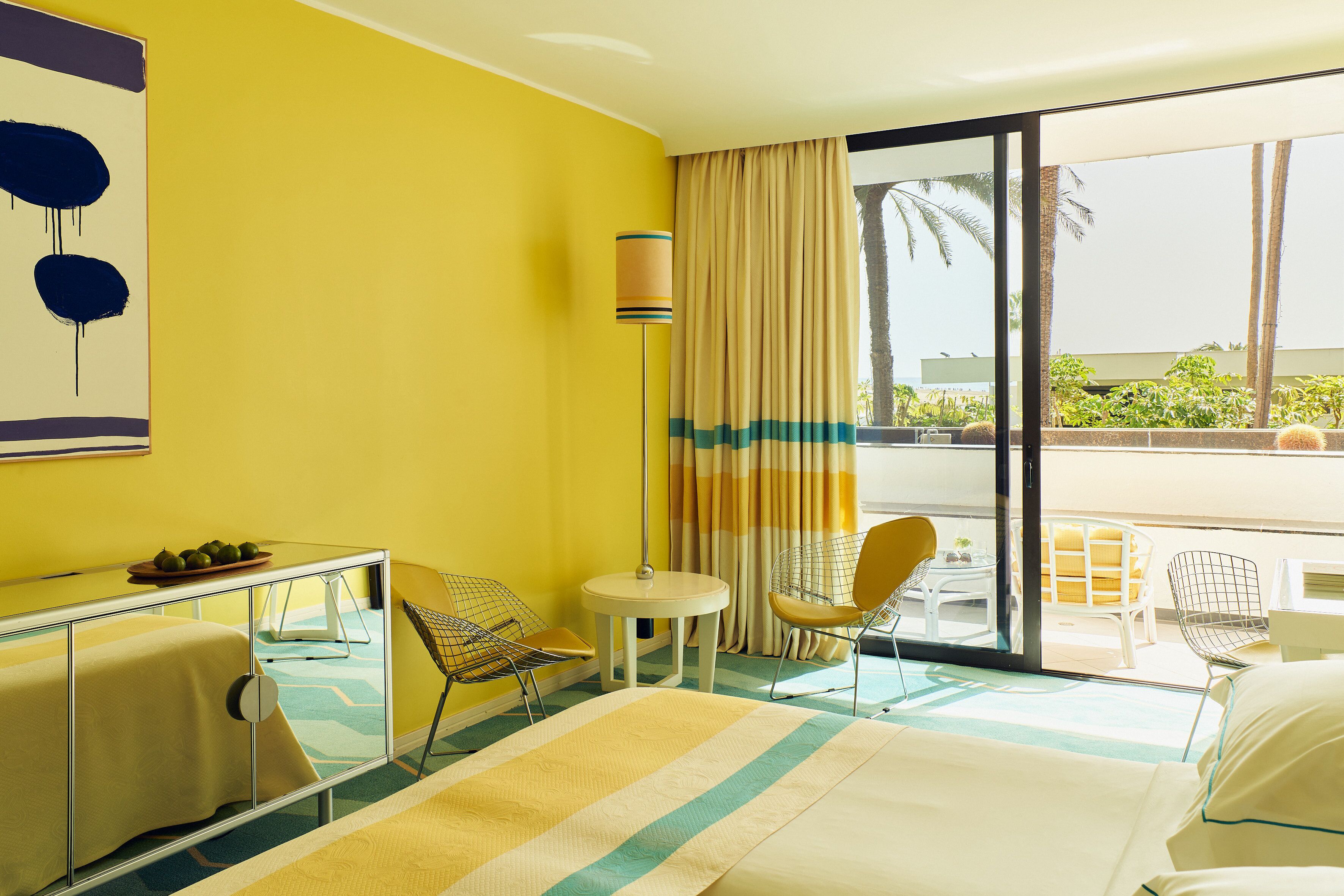 A hotel room with a bed, a desk with a chair, a small table with a chair, and a view of the outdoors.