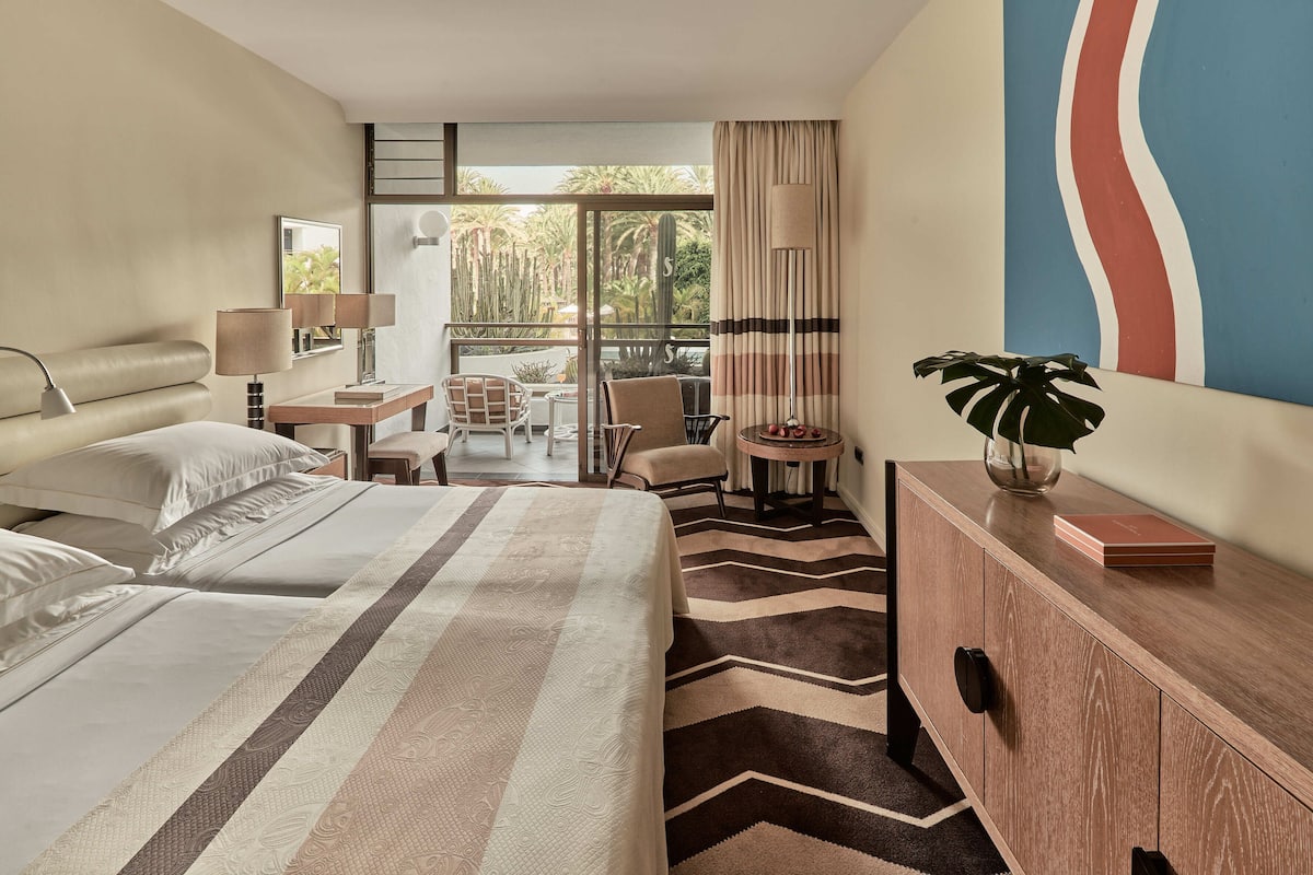 A hotel room with a bed, a desk, a chair, a small table with a plant, and a view of the outdoors.