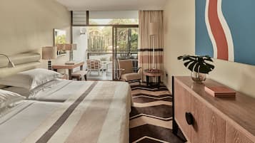 A hotel room with a bed, a desk, a chair, a small table with a plant, and a view of the outdoors.