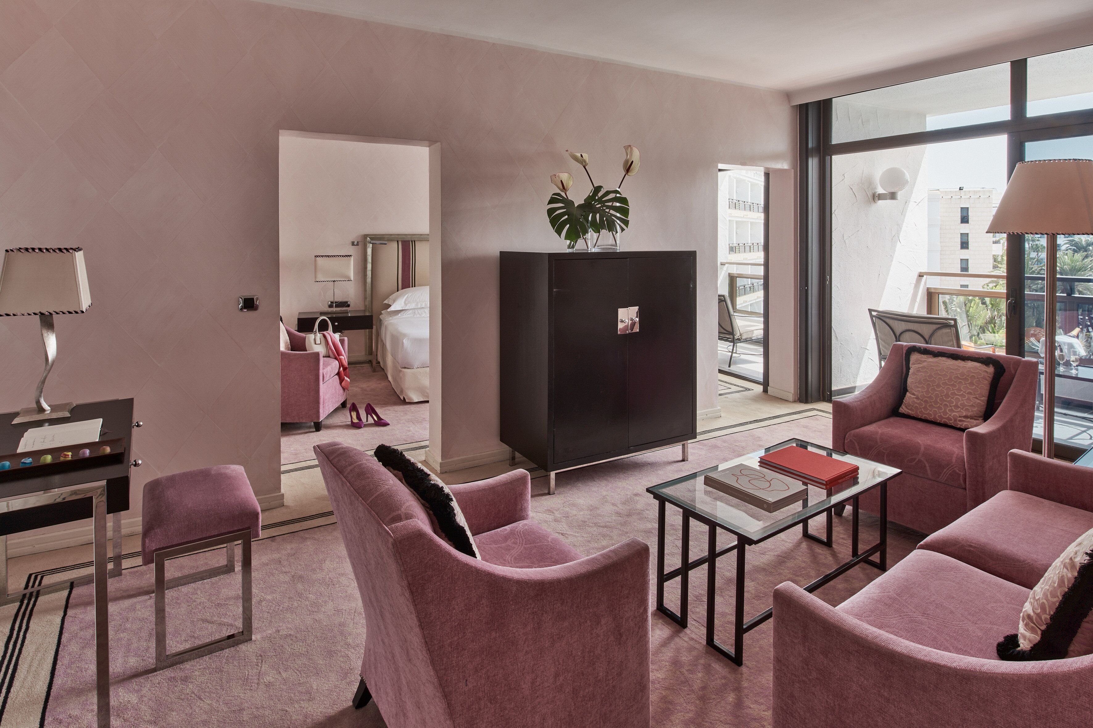 A modern hotel room with a pink color scheme, featuring a sofa, armchairs, a glass coffee table, and a black cabinet.