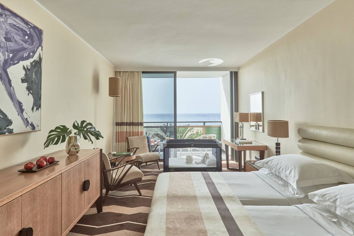 A hotel room with a bed, a desk, a chair, a TV, a plant, and a view of the ocean.