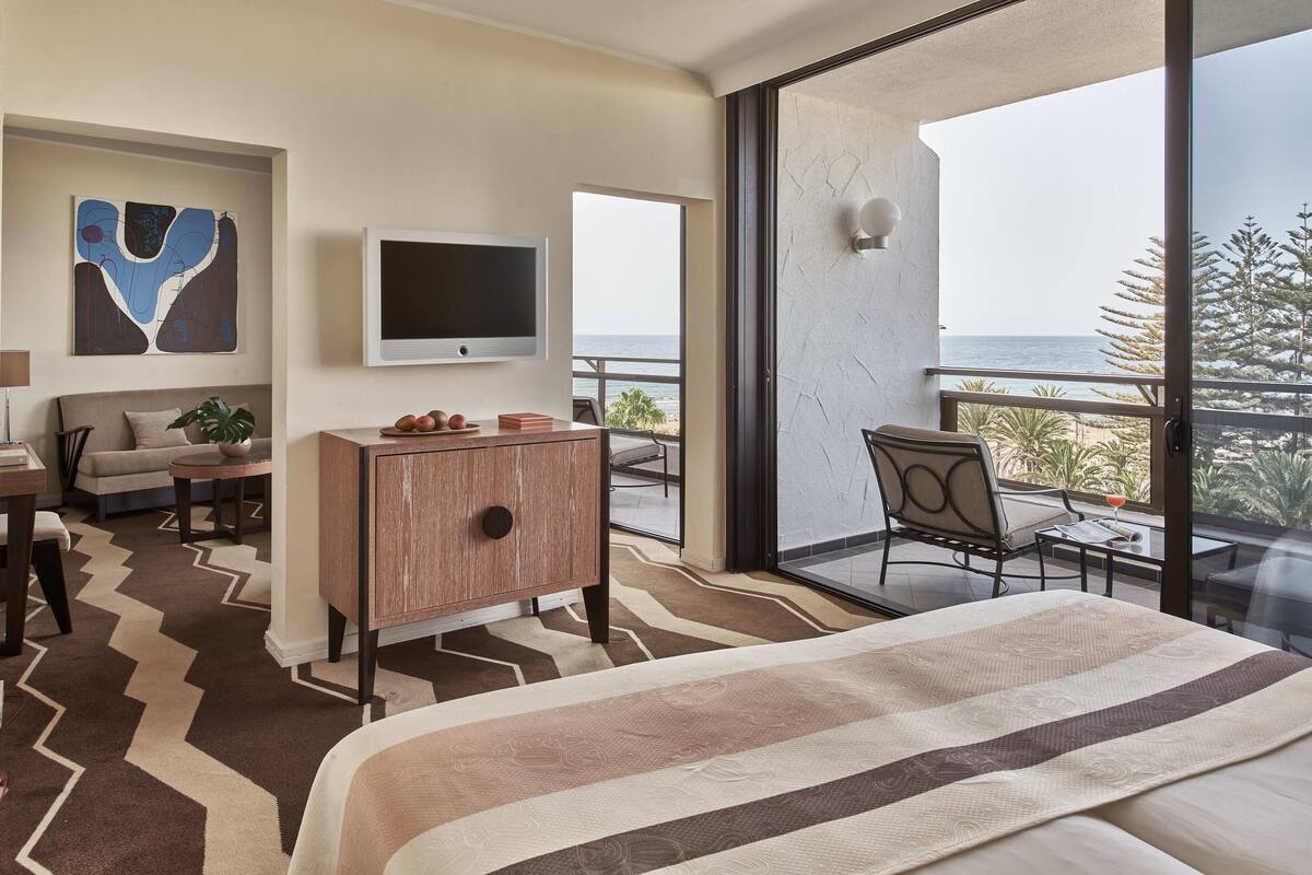 A hotel room with a large bed, a TV, a desk with a chair, a sofa, and a balcony with a view of the sea.