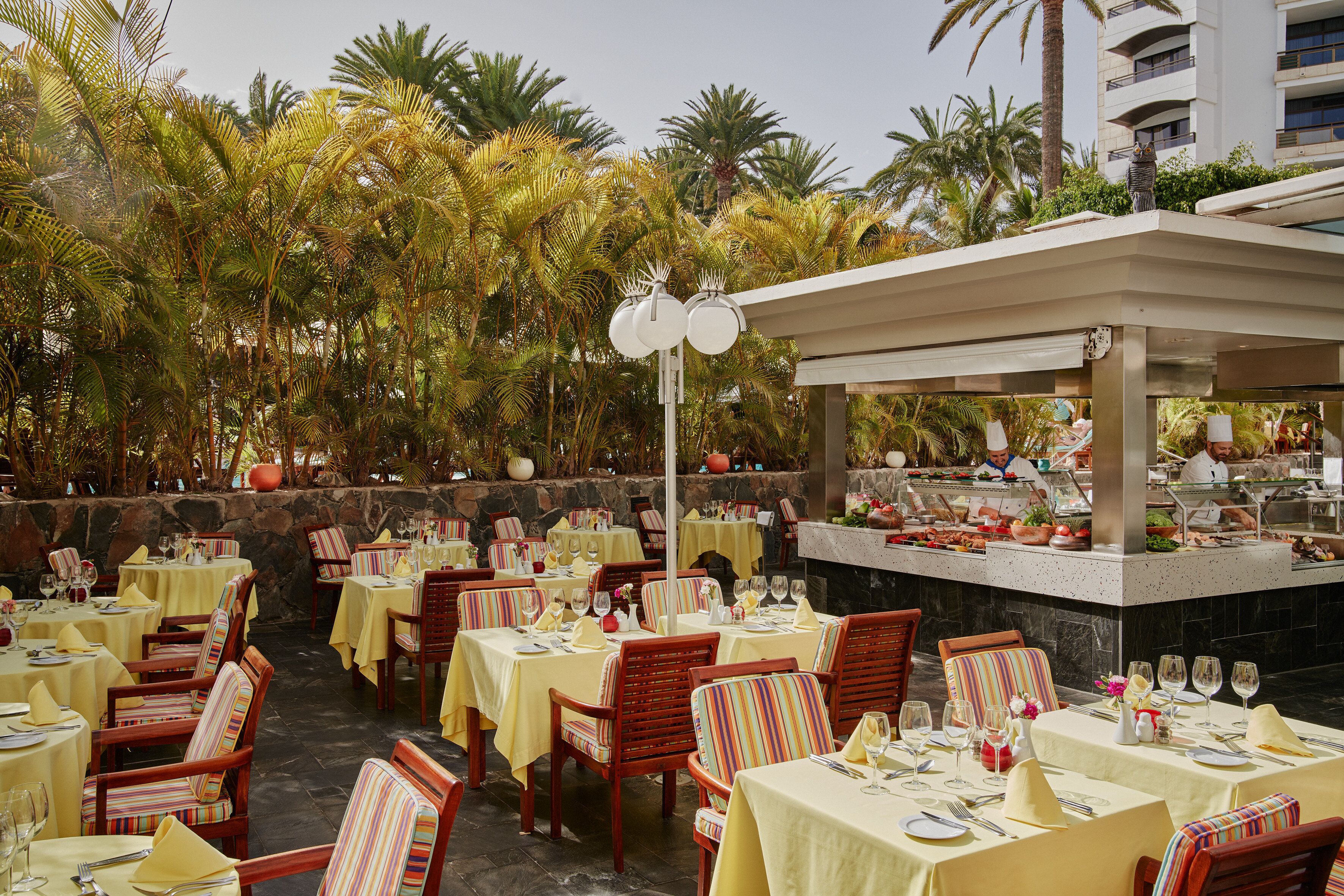 An outdoor dining area with tables and chairs, surrounded by palm trees and a stone wall.