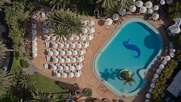 An aerial view of a resort with a swimming pool, sun loungers, umbrellas, and palm trees.