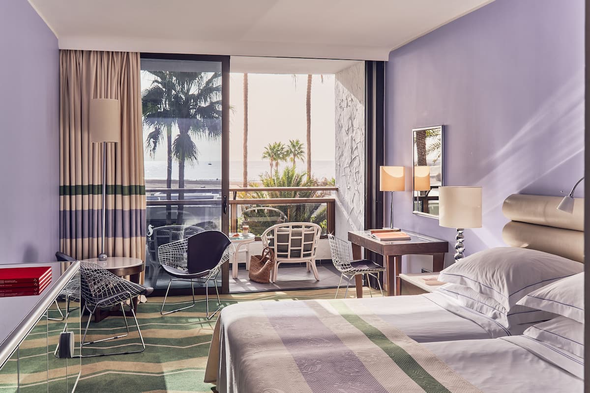 A hotel room with a bed, a desk, a chair, a small table, and a view of the ocean and palm trees.