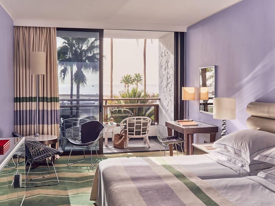 Habitación de hotel con cama, escritorio, silla, mesita y vista al océano y palmeras.