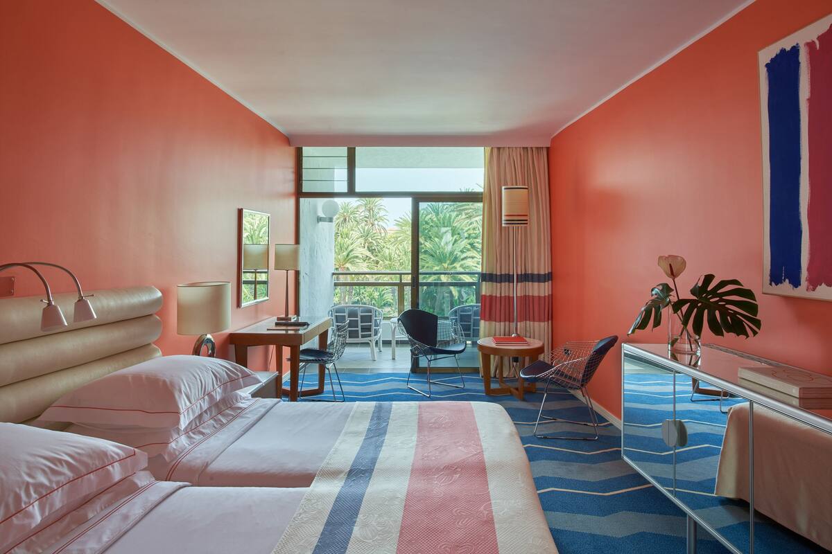 A hotel room with a large bed, a desk, a chair, and a balcony with a view of palm trees.