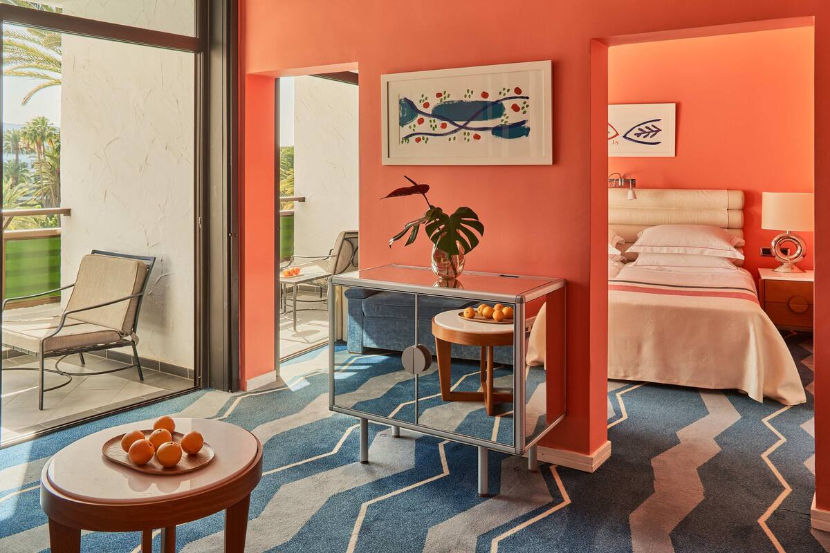 A hotel room with a bed, a bedside table, a chair, a small table with oranges, and a large window.