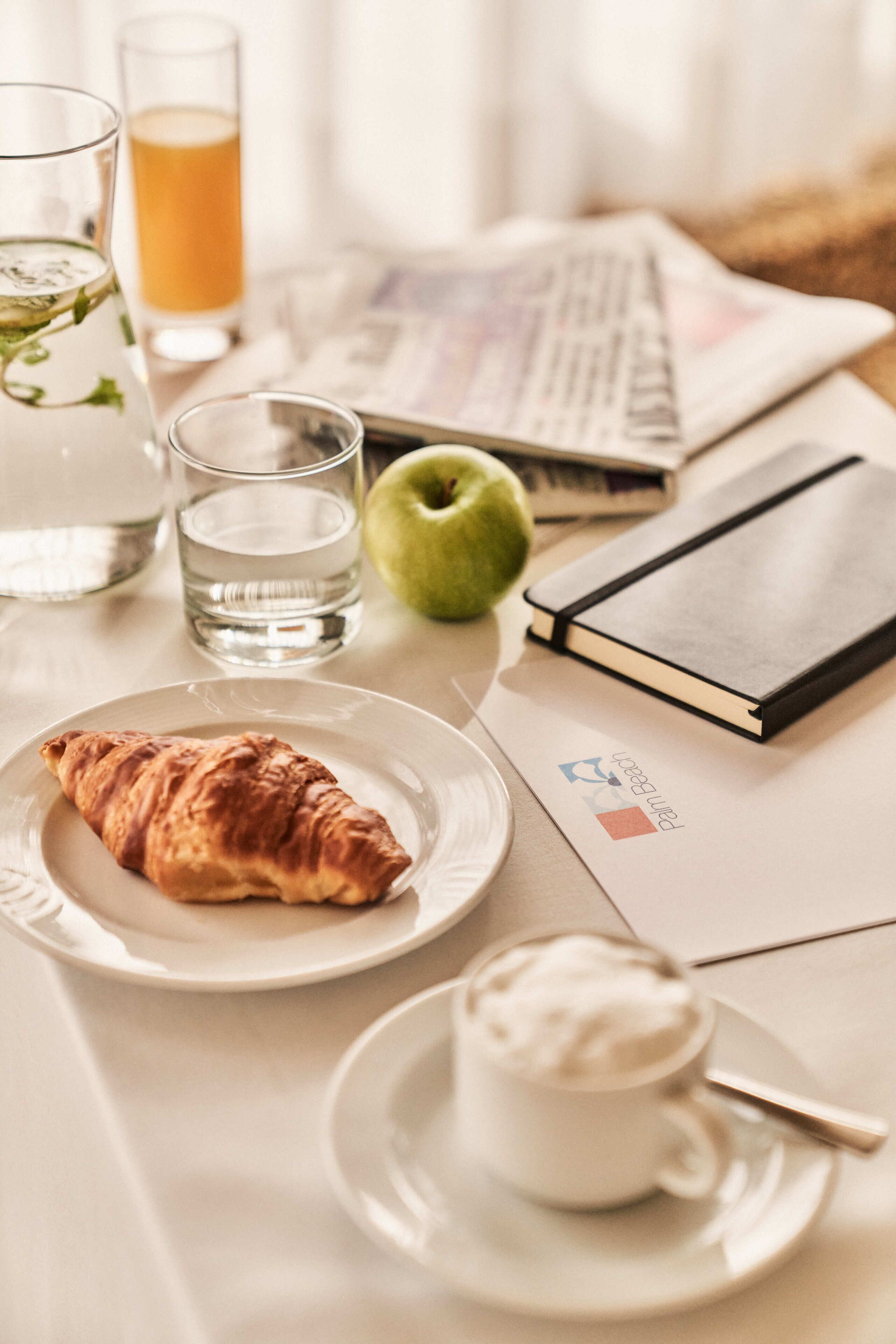 Une table mise pour le petit-déjeuner avec un croissant, un verre d’eau, un verre de jus d’orange, une pomme verte, un carnet et une tasse de café.