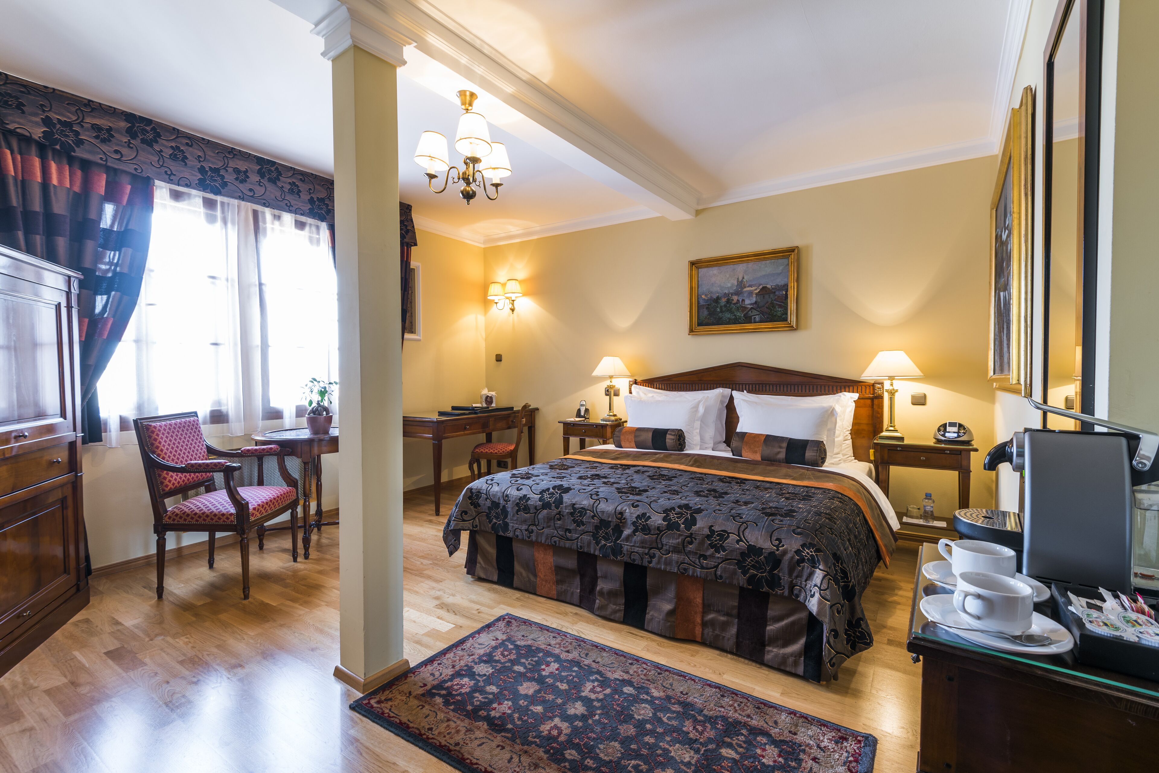 Quarto de hotel com uma cama grande, uma escrivaninha com cadeira, uma mesinha e uma mesa de cabeceira com luminária.