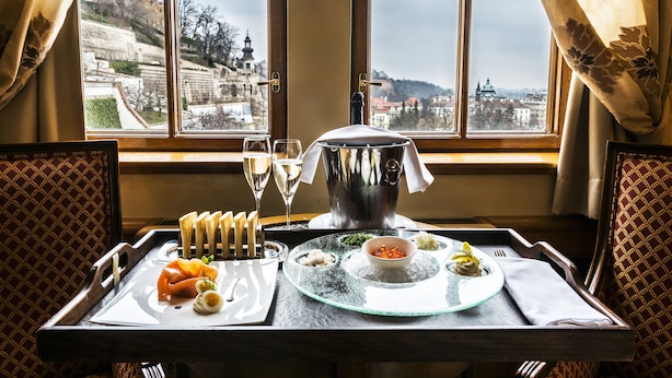 A dining table with a view of a cityscape through a window, a bottle of champagne, and a tray with food items. - Golden Well Hotel (Prague)