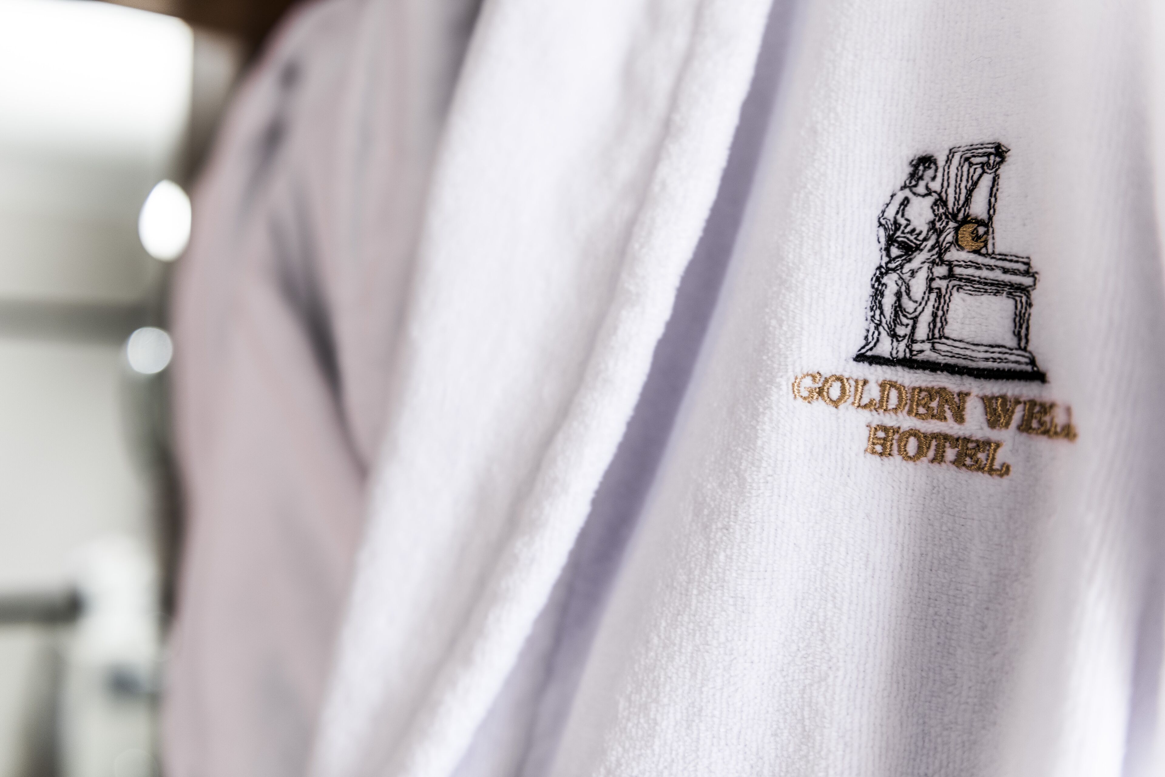 A white garment with an embroidered logo of a person standing next to a pedestal, and the text "GOLDEN WELL HOTEL" below it.