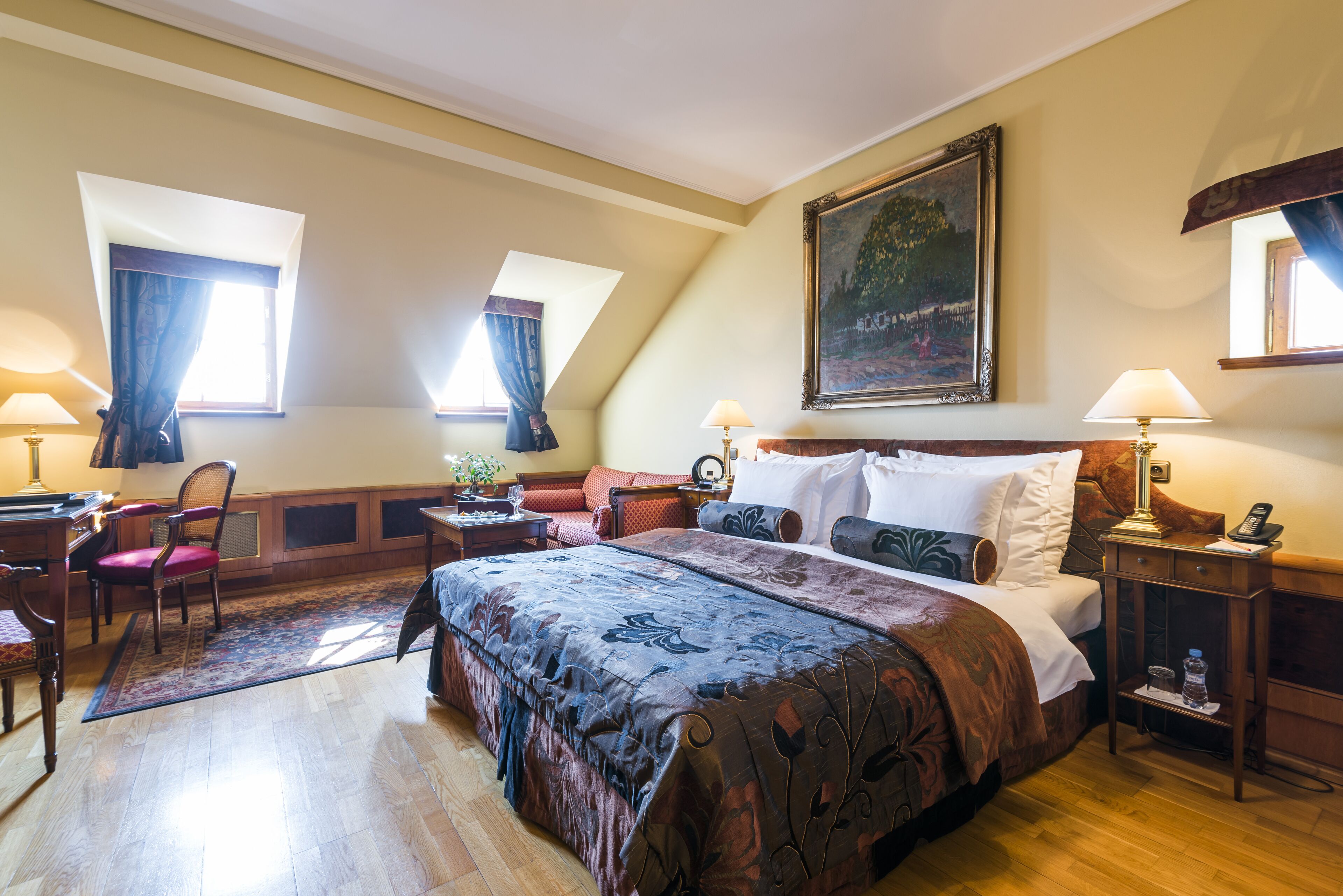 A hotel room with a large bed, a desk with a chair, a small table with a lamp, and a painting on the wall.