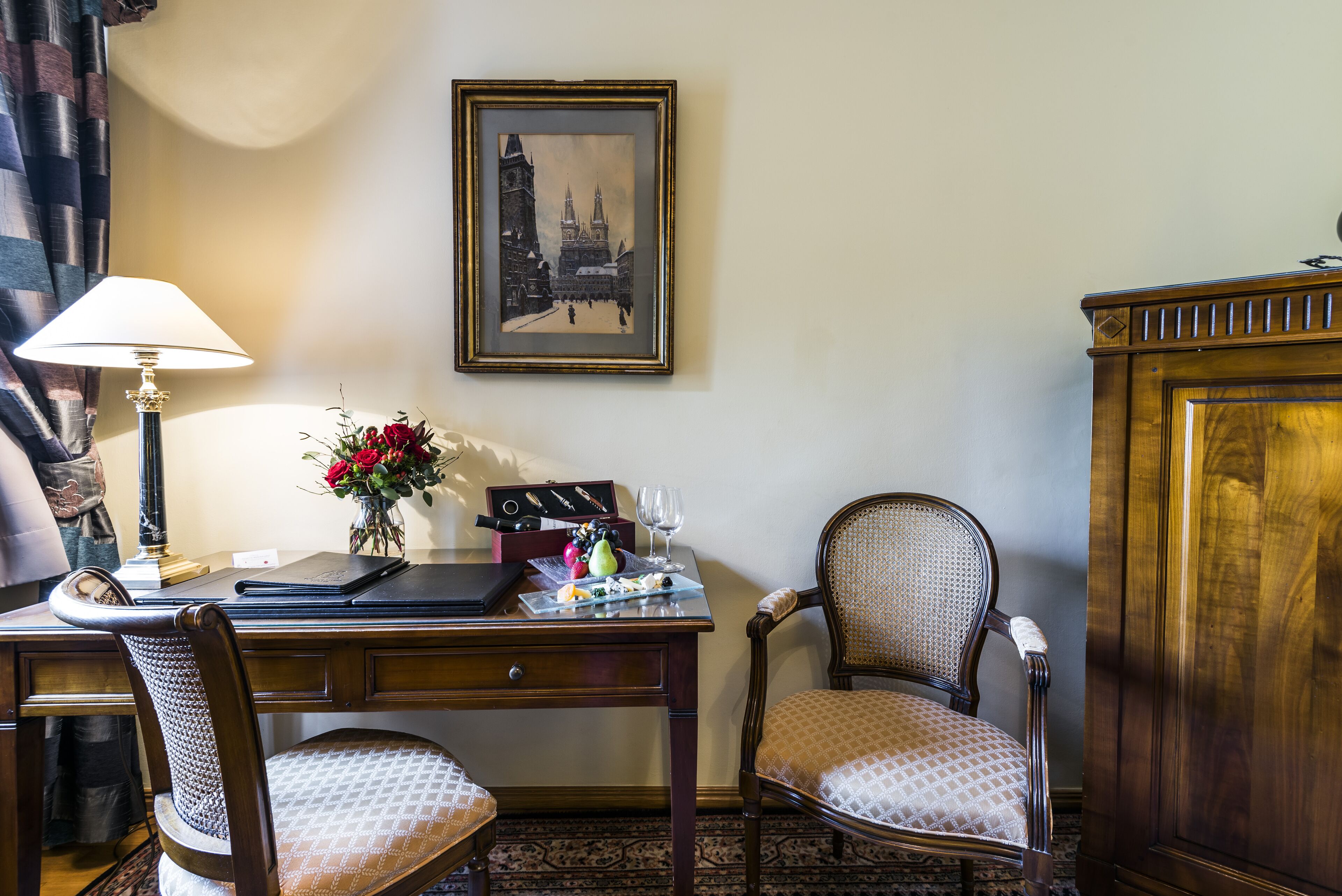A hotel room with a wooden desk, two chairs, a lamp, a framed picture on the wall, and a cabinet.