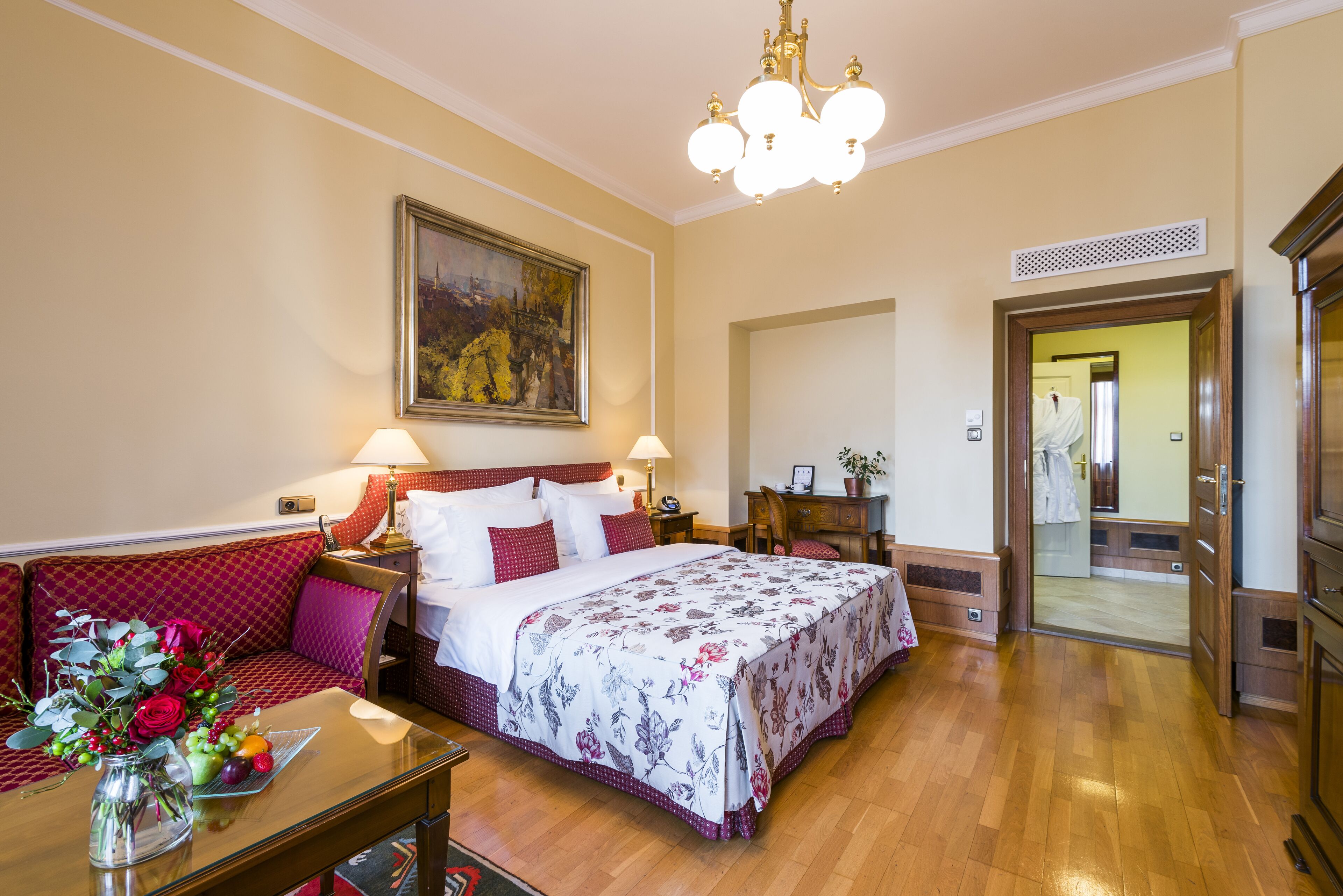 A hotel room with a large bed, a desk, a chair, a sofa, a chandelier, and a painting on the wall.