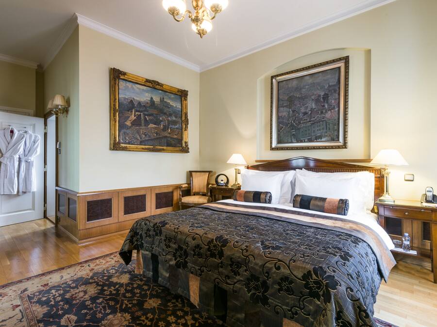 A hotel room with a large bed, two paintings on the walls, a wooden nightstand, a desk, and a robe rack.