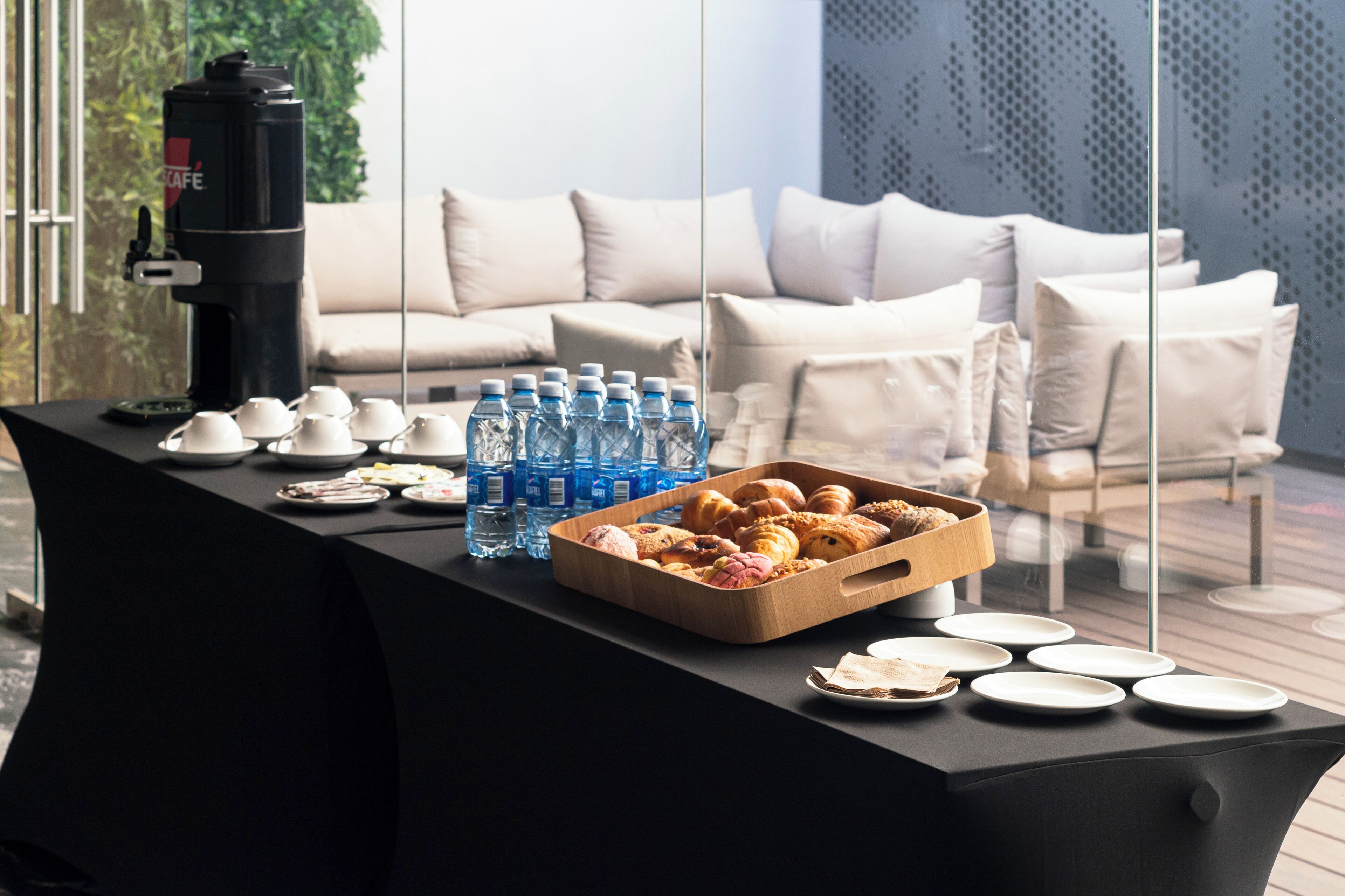 Una mesa con una máquina de café, botellas de agua, pasteles y platos, frente a un interior moderno con un sofá y paredes de vidrio.