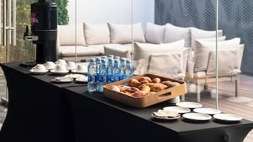Una mesa con una máquina de café, botellas de agua, pasteles y platos, frente a un interior moderno con un sofá y paredes de vidrio.