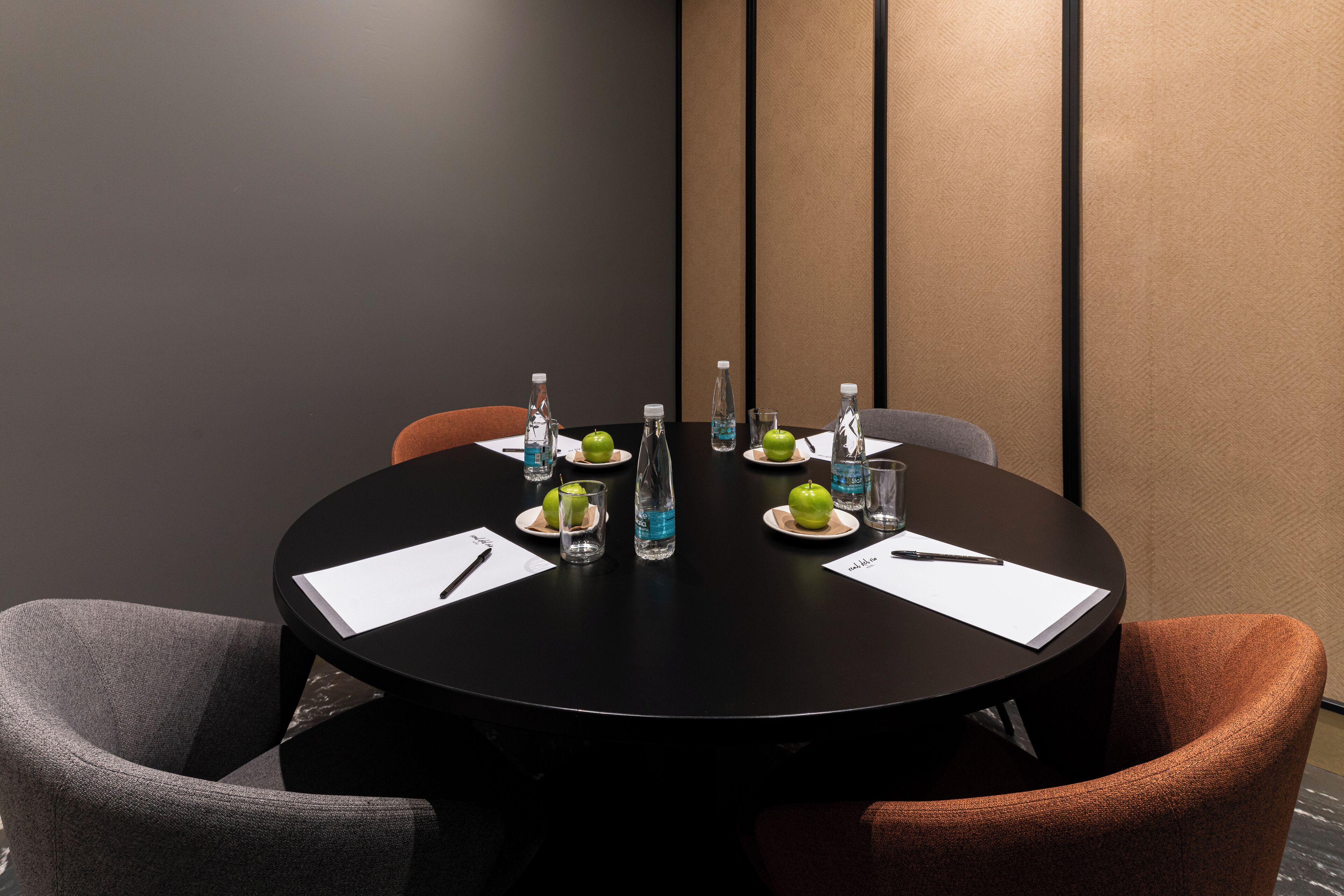 A round table with a black surface, surrounded by four chairs, two grey and two orange. There are two green apples, two bottles of water, and two glasses on the table.