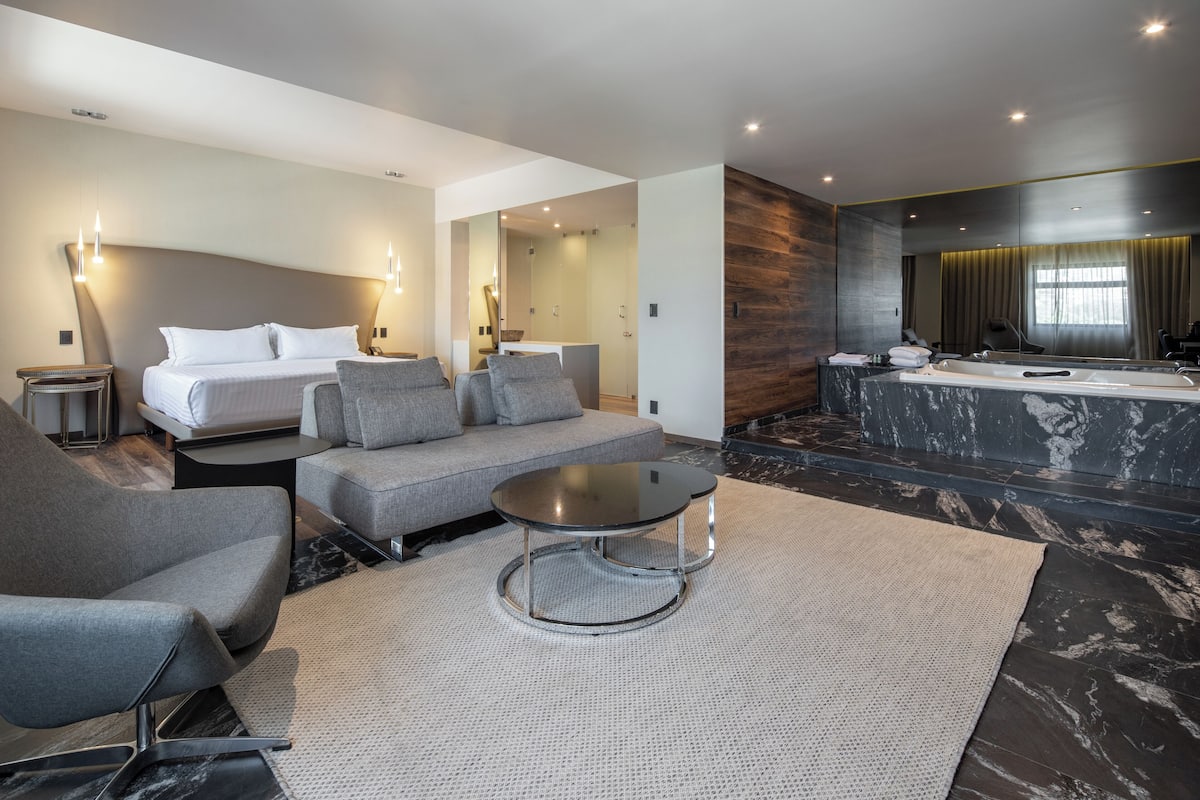 A modern hotel room with a bed, sofa, armchair, and a small round table.