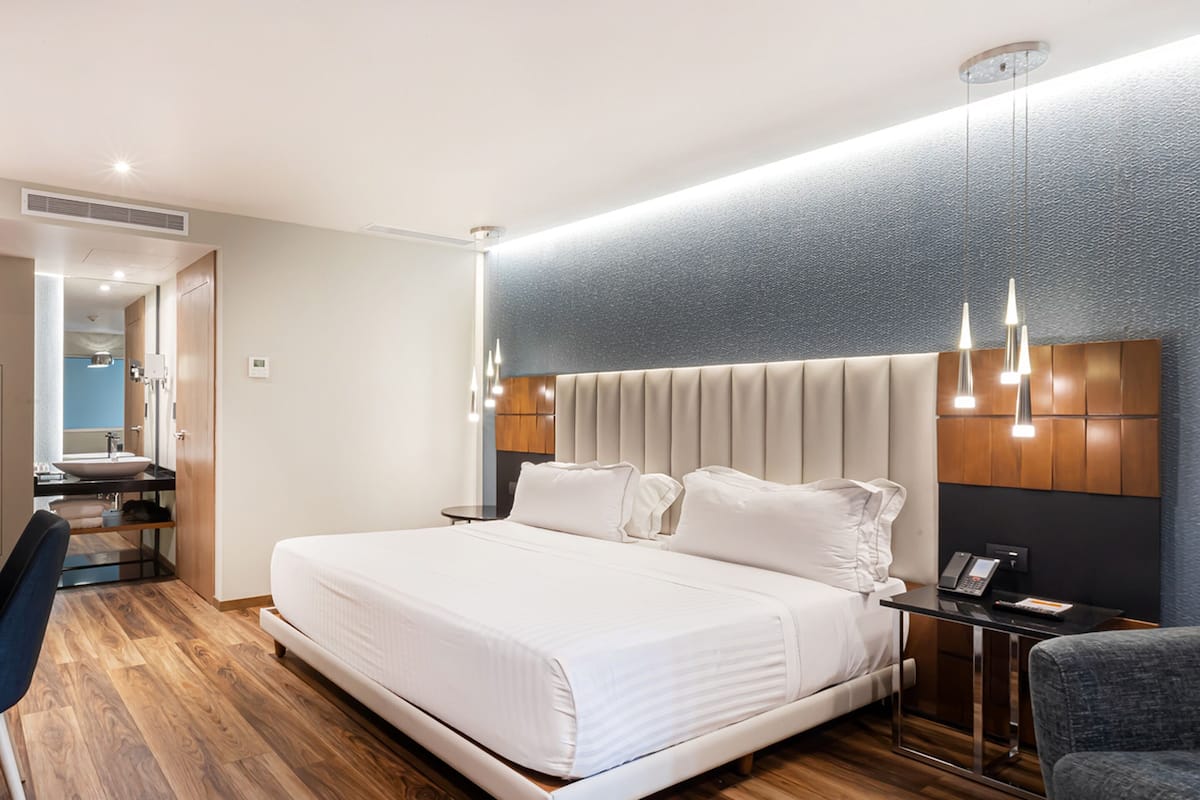 A modern hotel room with a large bed, a desk with a sink, and a mirror.