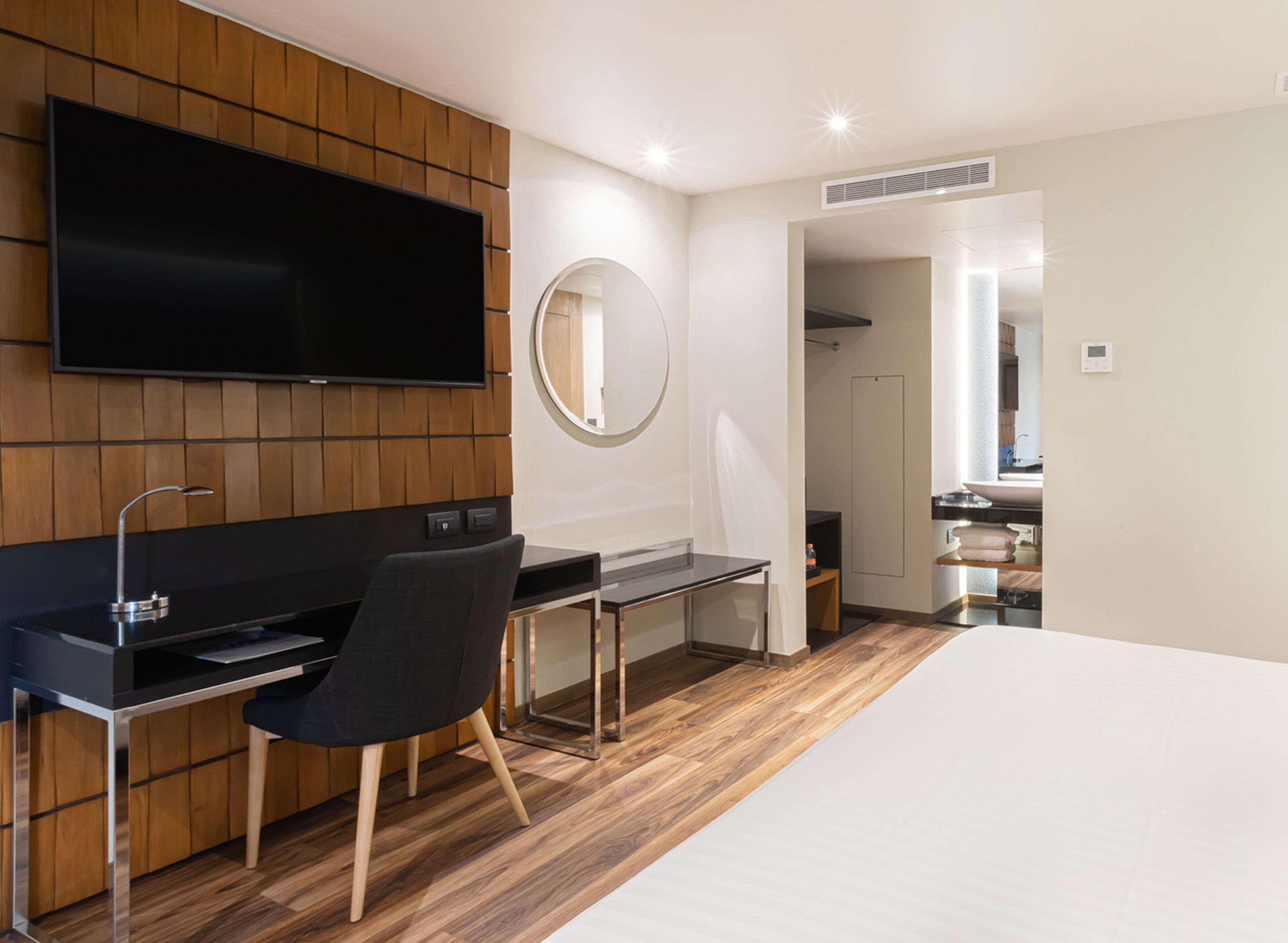 A modern hotel room with a flat-screen TV mounted on a wooden panel wall, a round mirror, a desk with a chair, and a large bed.