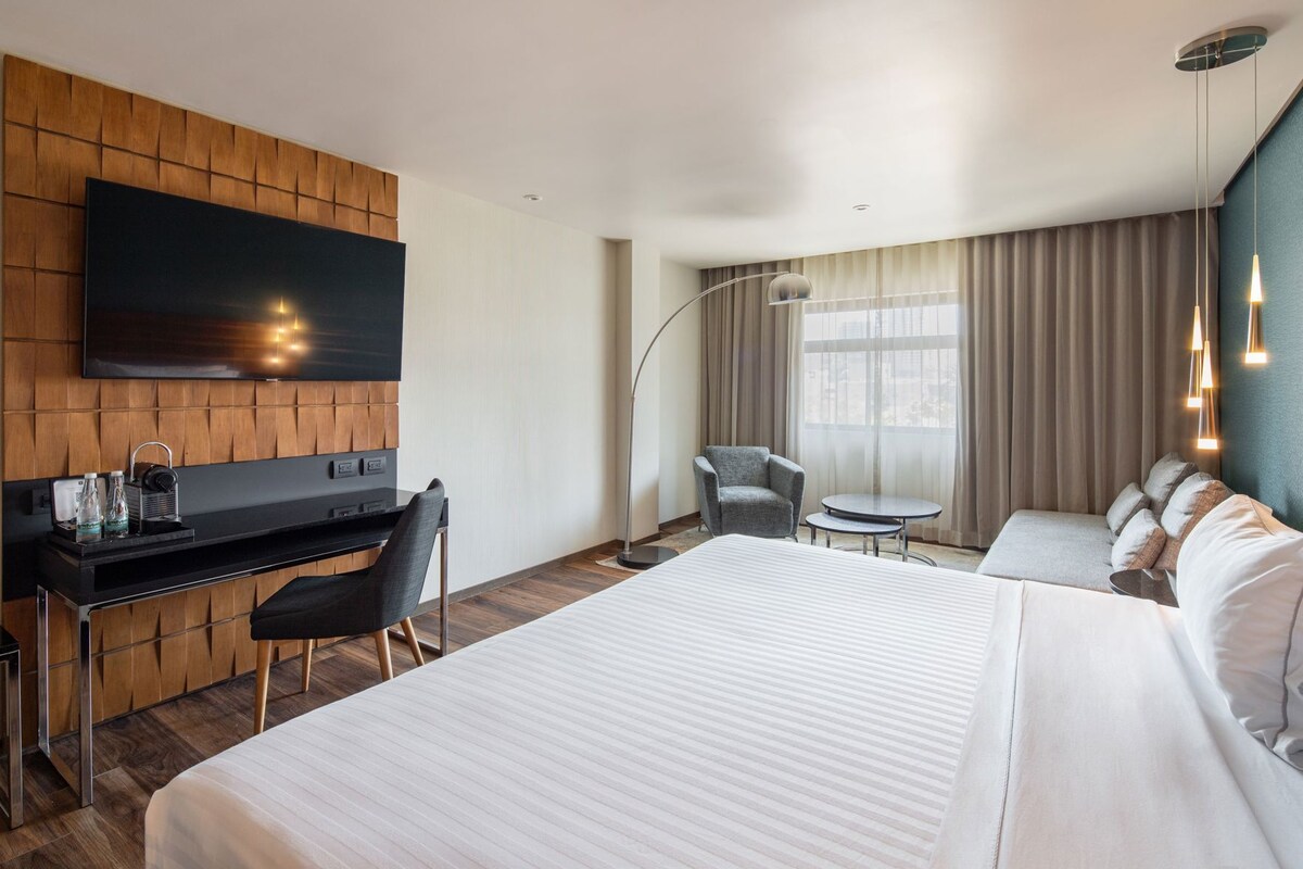 A hotel room with a large bed, a desk with a chair, a television, and a seating area with a chair and a small table.