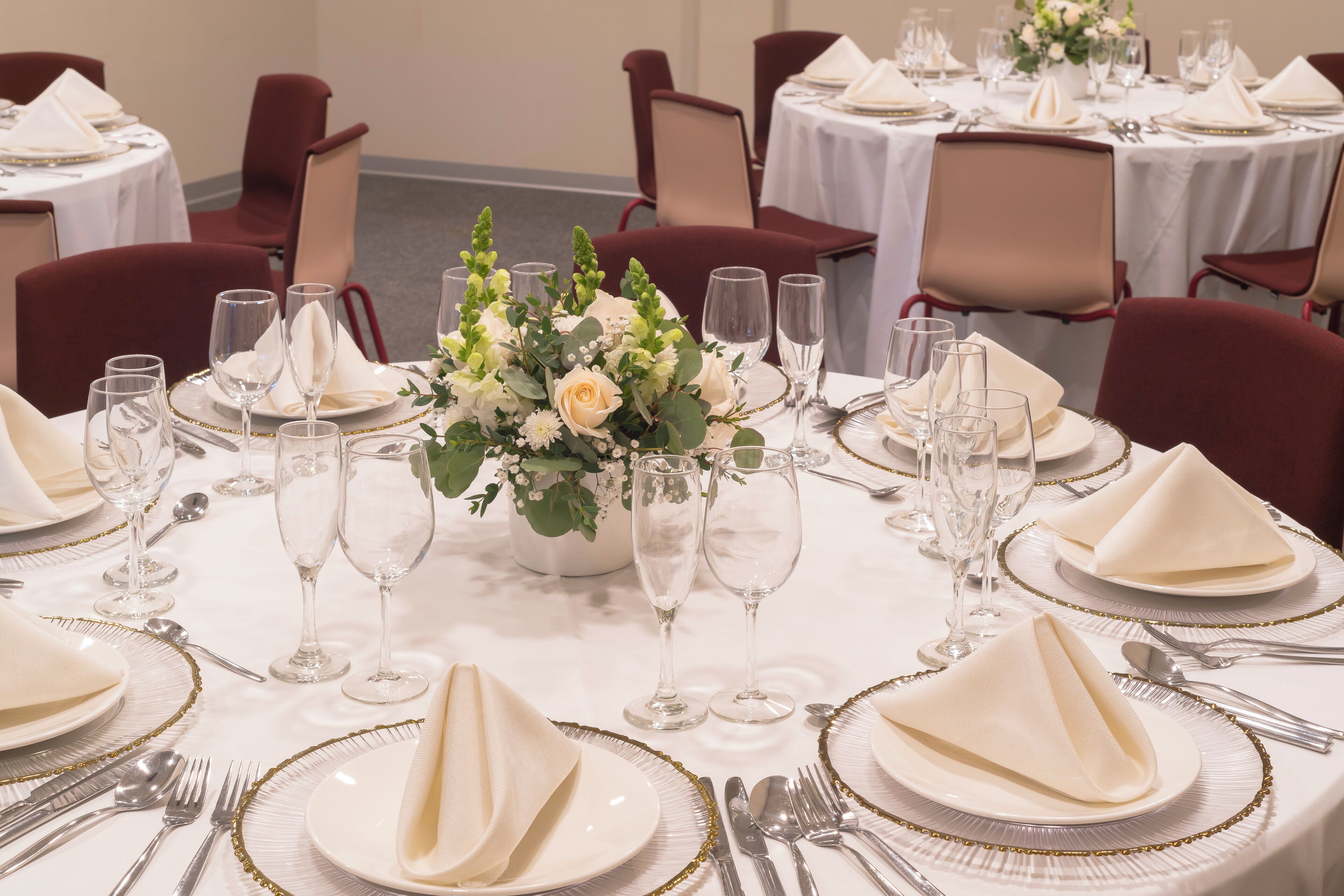 A banquet hall with round tables set for a formal event, complete with white tablecloths, gold-trimmed place settings, and floral centerpieces.