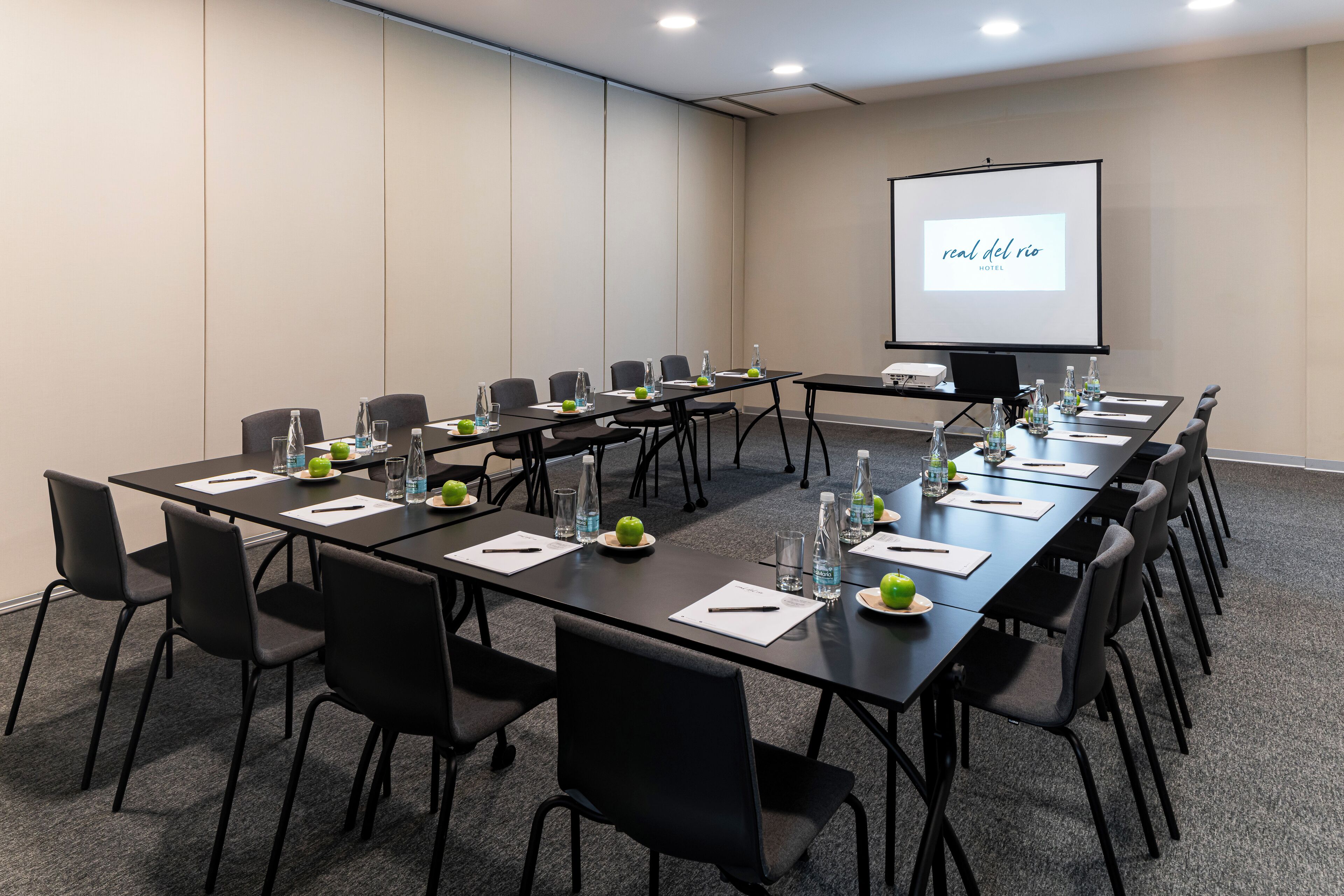 A conference room with long tables, chairs, water bottles, and a projector screen displaying "real del rio hotel".