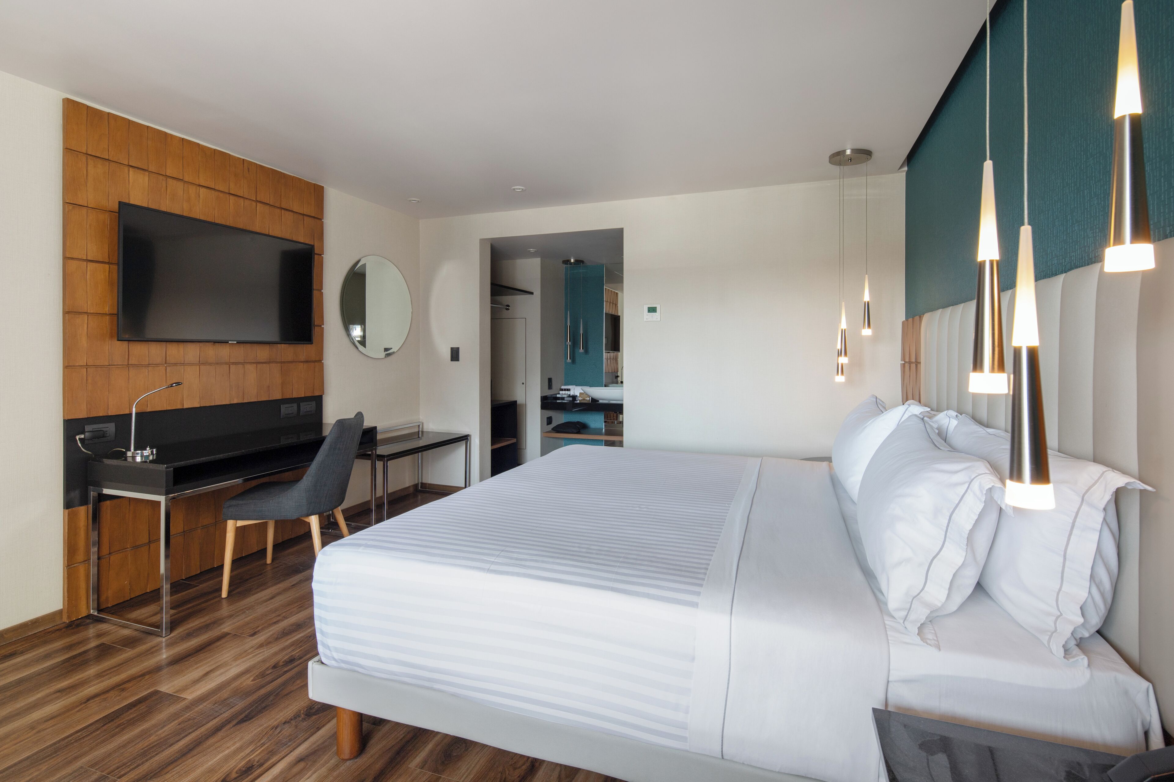 Una moderna camera d'albergo con un letto grande, una scrivania con una sedia, una TV a schermo piatto fissata al muro e lampade a sospensione sopra il letto.