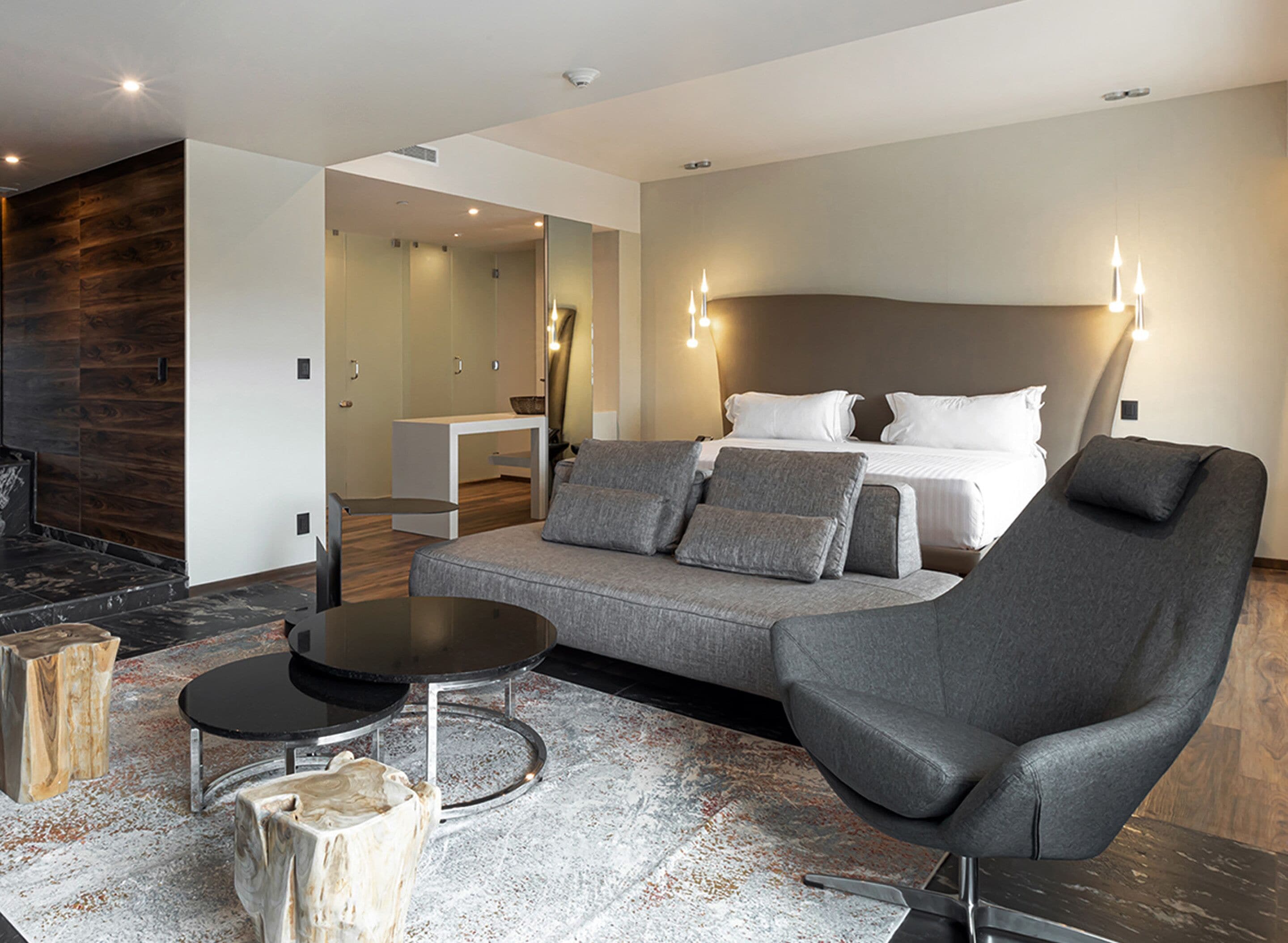 Ein modernes Hotelzimmer mit einem großen Bett, einem grauen Sofa und einem Sessel. Es gibt einen kleinen Tisch und einen Couchtisch aus Holz. Der Boden ist aus Holz und über dem Bett befindet sich ein großer Spiegel.
